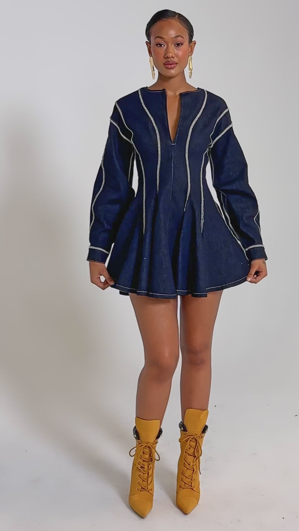 A video of a woman wearing the PLAYDATE DENIM MINI DRESS, a navy blue long-sleeved fit and flare mini dress with white seam detailing and a front keyhole cutout, styled with mustard yellow lace-up ankle boots.