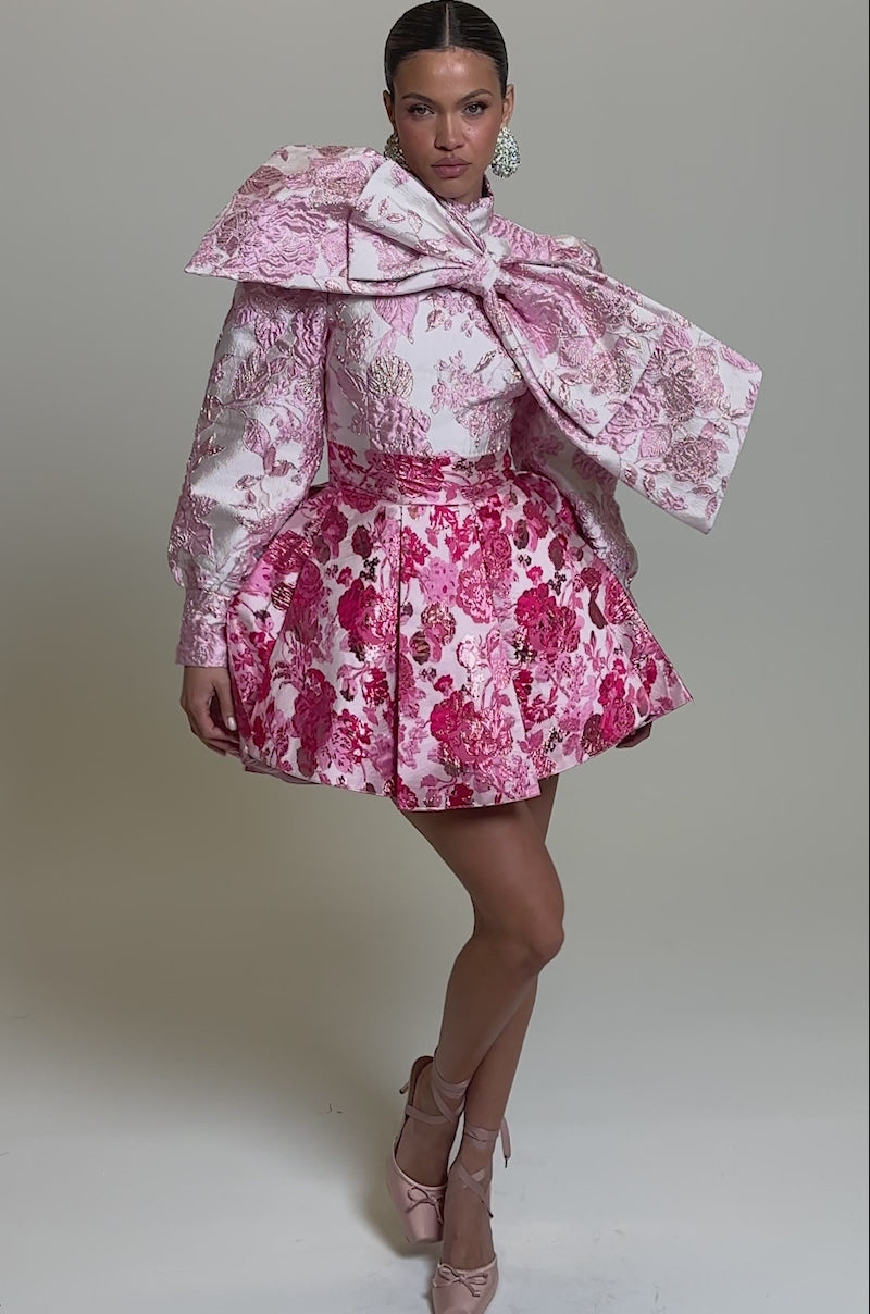Video of woman modeling the RENAISSANCE TWO-TONE BROCADE MINI DRESS IN PINK. The dress features pink and white floral jacquard, a statement neck bow, voluminous long sleeves, and a flared mini skirt.