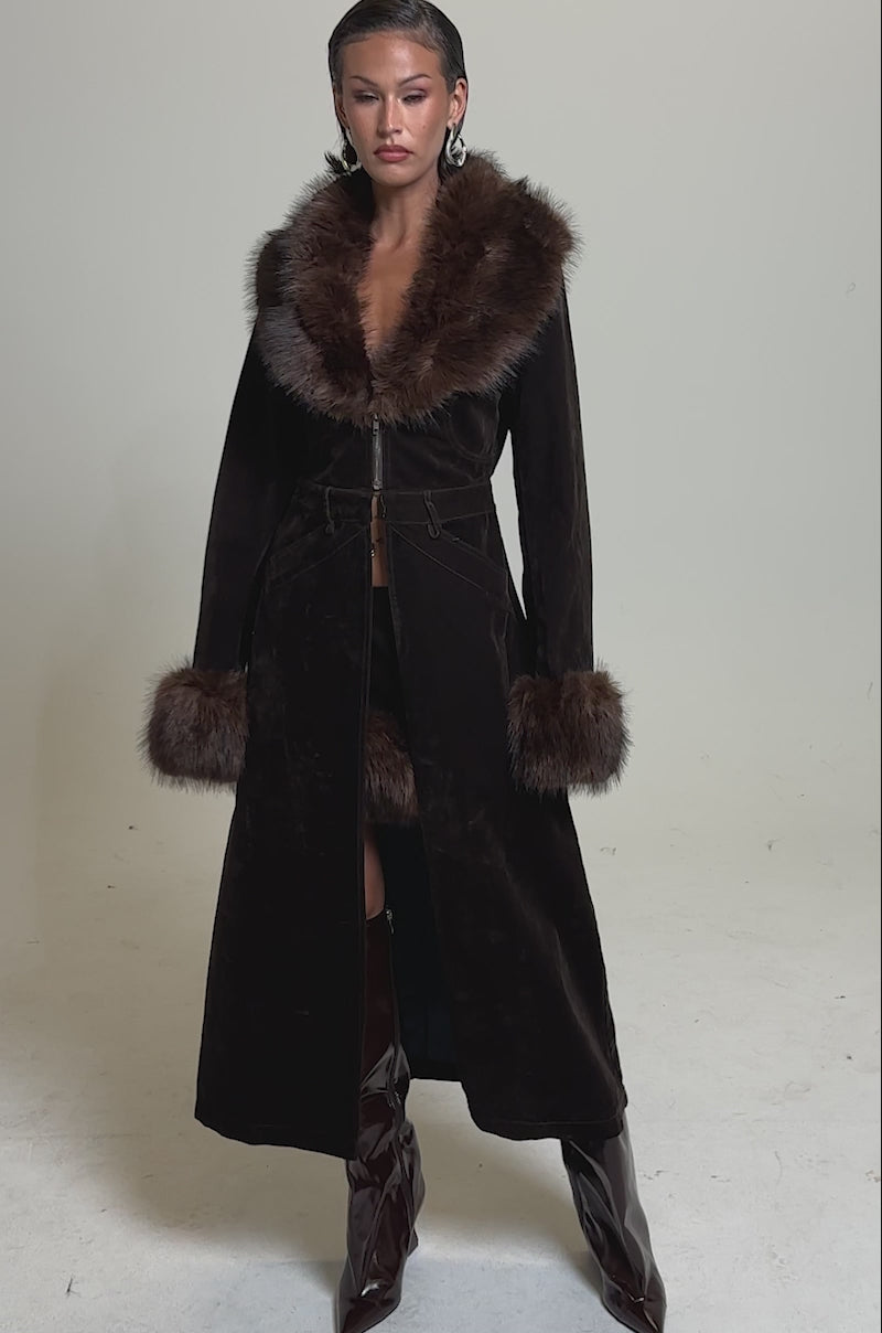 A video of a woman in the WILD SIDE VELVET DENIM TRENCH with a luxe faux fur collar and cuffs, a matching brown mini skirt underneath, and knee-high dark boots stands against a plain light background.