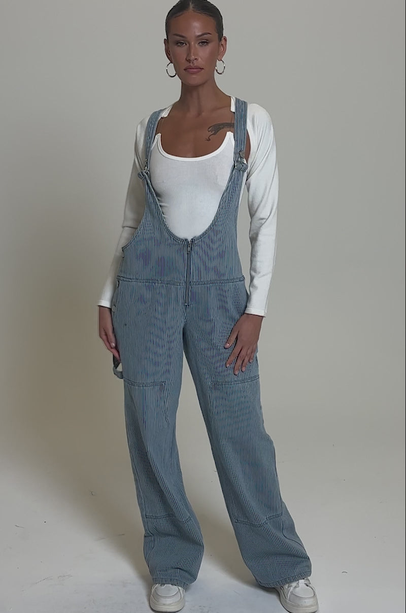 Video of woman modeling the HAMPTONS STRIPED CARPENTER OVERALLS over a fitted white long-sleeve shirt and white sneakers, hair pulled back with large hoop earrings