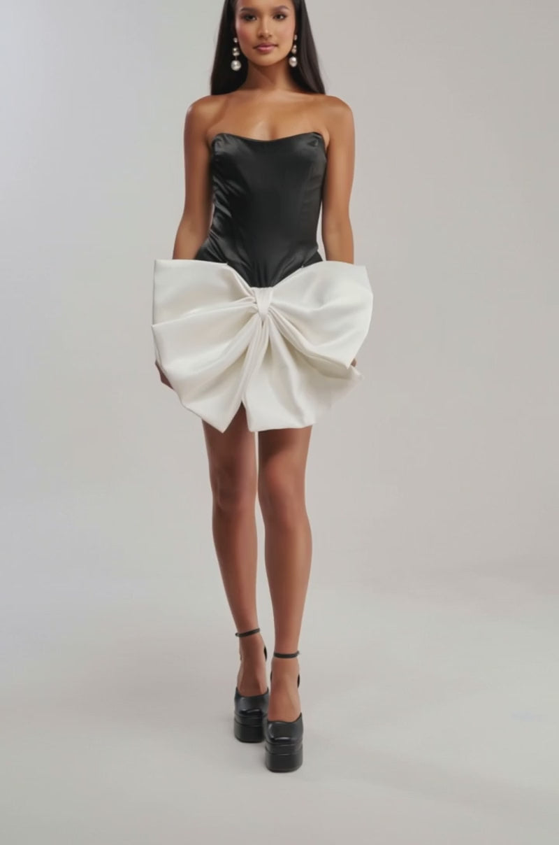 Video of woman modeling the SITTING PRETTY BOW MINI DRESS—a strapless black satin mini dress with an oversized white waist bow—paired with black platform heels. She has long, straight hair and stands against a plain light gray background.
