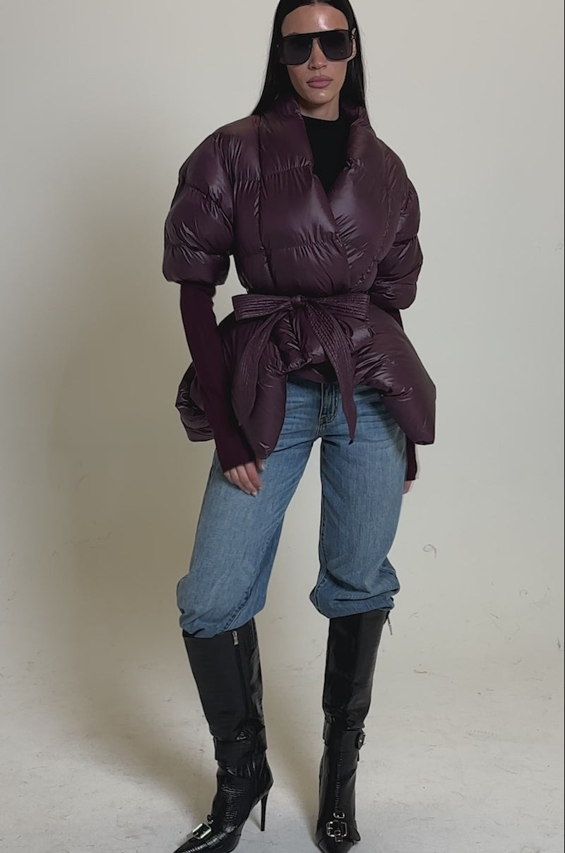 A video of a woman in the RIB SLEEVE PEPLUM PUFFER IN PURPLE, blue cuffed jeans, black stiletto boots, a black turtleneck, and sunglasses poses against a plain gray background.
