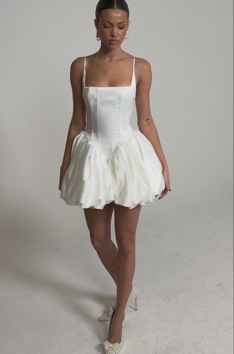 A video of a woman wearing the JULIET SATIN MINI DRESS IN IVORY with a corset bodice and bubble skirt, styled with white high heels, standing against a plain background.