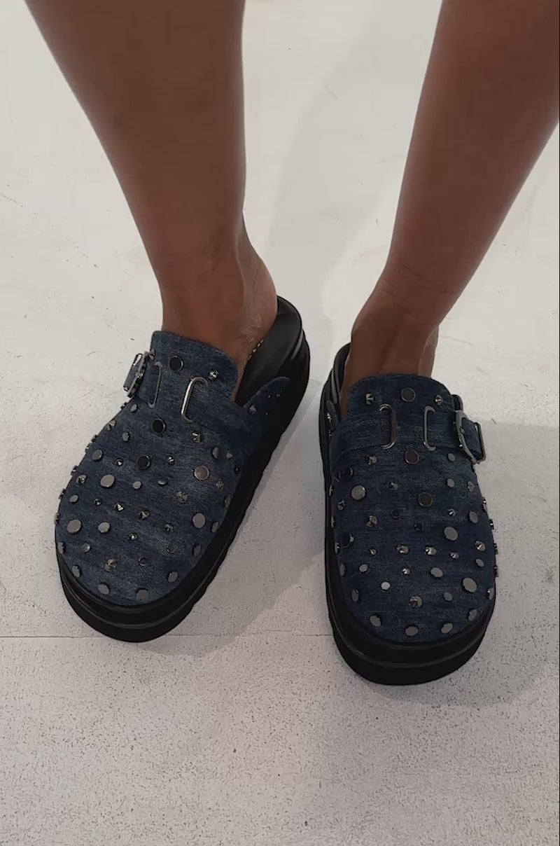 A video of a model wearing the AKIRA GOLD BILLIANNA DENIM FLAT CLOG WITH HARDWARE—blue denim slip-on clogs adorned with silver studs and set on black soles.