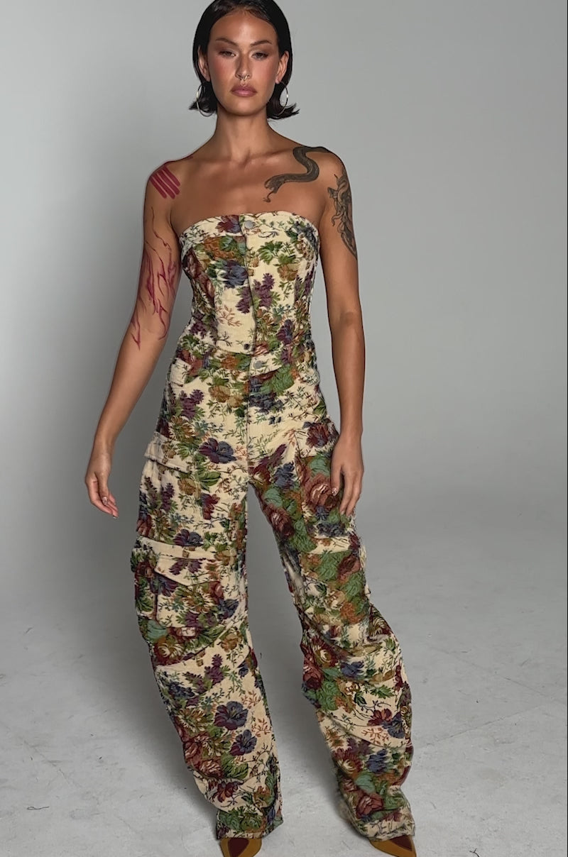 A video of a person in the IN HER ELEMENT FLORAL WOVEN JUMPSUIT, featuring a strapless design, cargo pockets, and wide legs. With short hair and visible tattoos on one arm, they pose with hands in pockets against a light gray background.