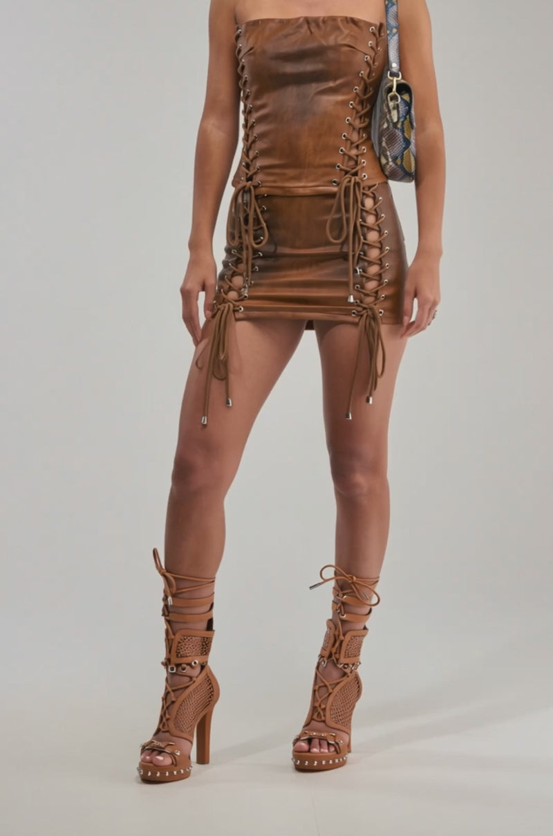 A video of a woman wearing the LOCK ME UP LACE UP MINI SKIRT in brown leather with lace-up sides, paired with matching heels and a patterned shoulder bag, standing against a plain light background.