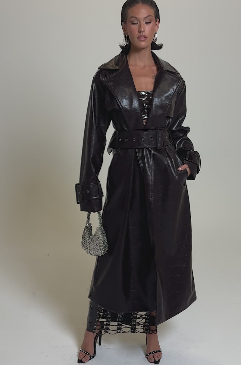 A video of a woman in the EMBOSSED CROC TRENCH, a long, belted dark brown crocodile-pattern coat. She holds a silver handbag, wears high-heeled sandals, and reveals a lace-up skirt beneath the coat.