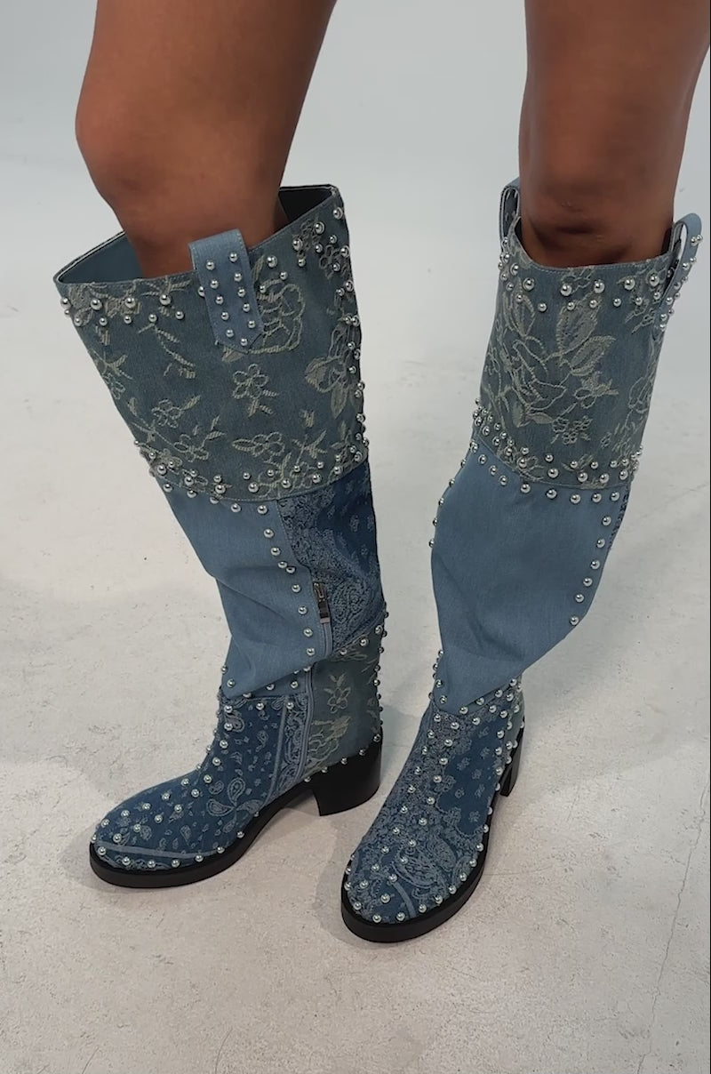 Video of woman modeling the AZALEA WANG LUVIENNE DENIM PATCHWORK STUDDED BLOCK BOOT—knee-high blue denim cowboy boots with silver studs and floral embroidery—paired with a blue skirt featuring 3D floral embellishments.