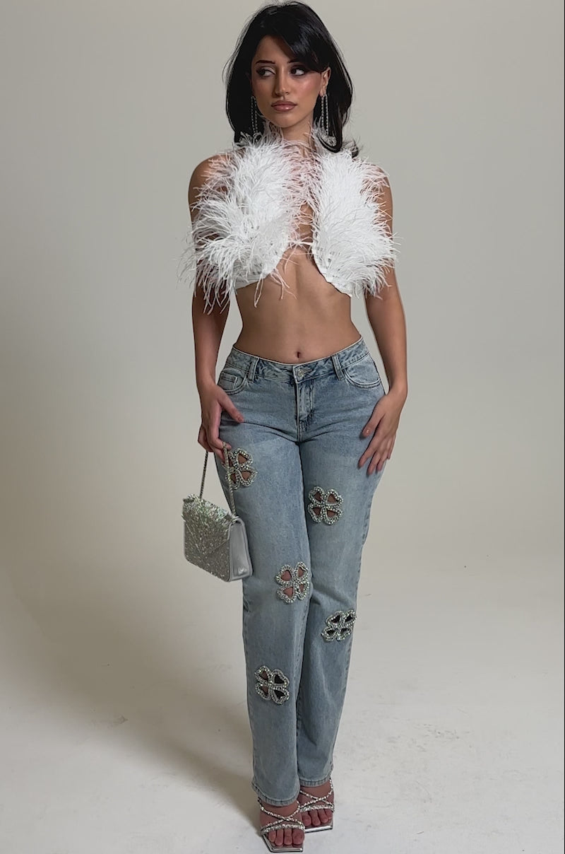 A video of a woman in the DIVINE FEMININE ENERGY RHINESTONE DETAIL JEANS, a white feathered crop top, silver sandals, and holding a small silver purse against a plain gray background.