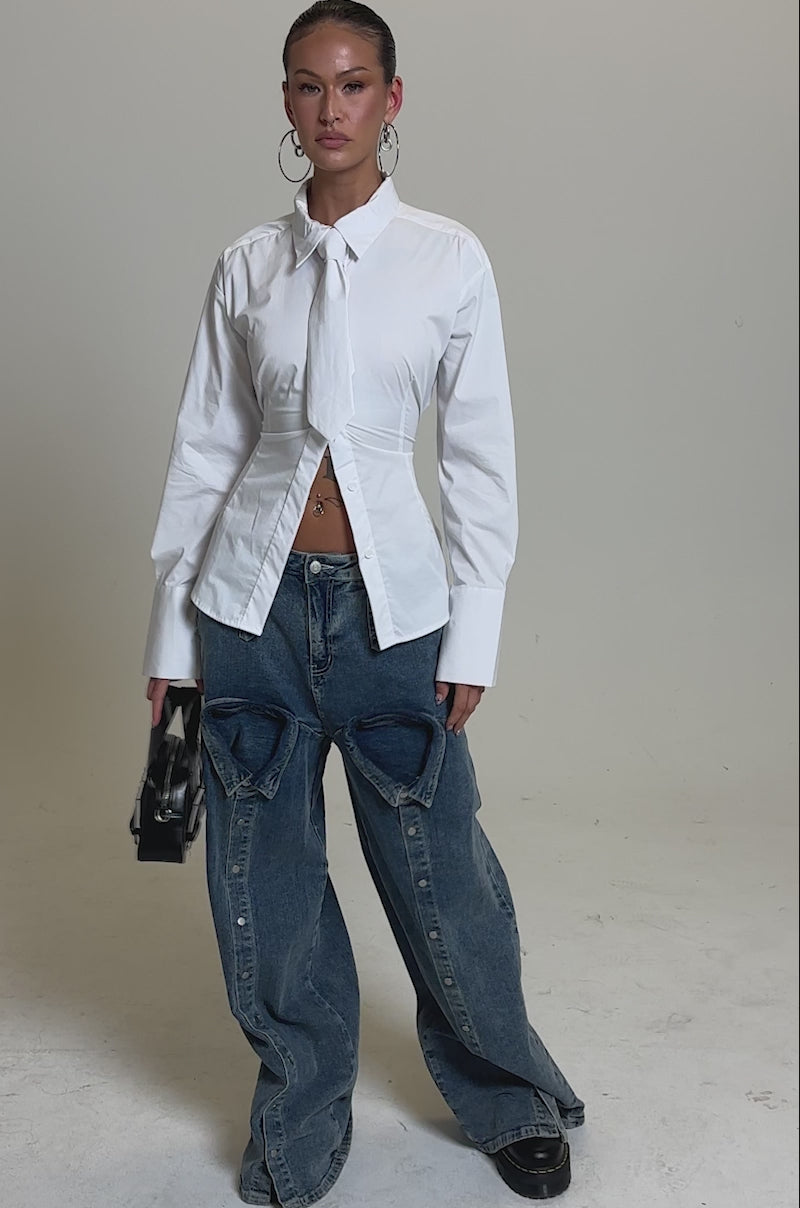 A video of a woman standing holding a small black handbag, wearing the BUSINESS BUTTON DOWN in white and oversized blue jeans with large hip cutouts, against a plain light gray background.