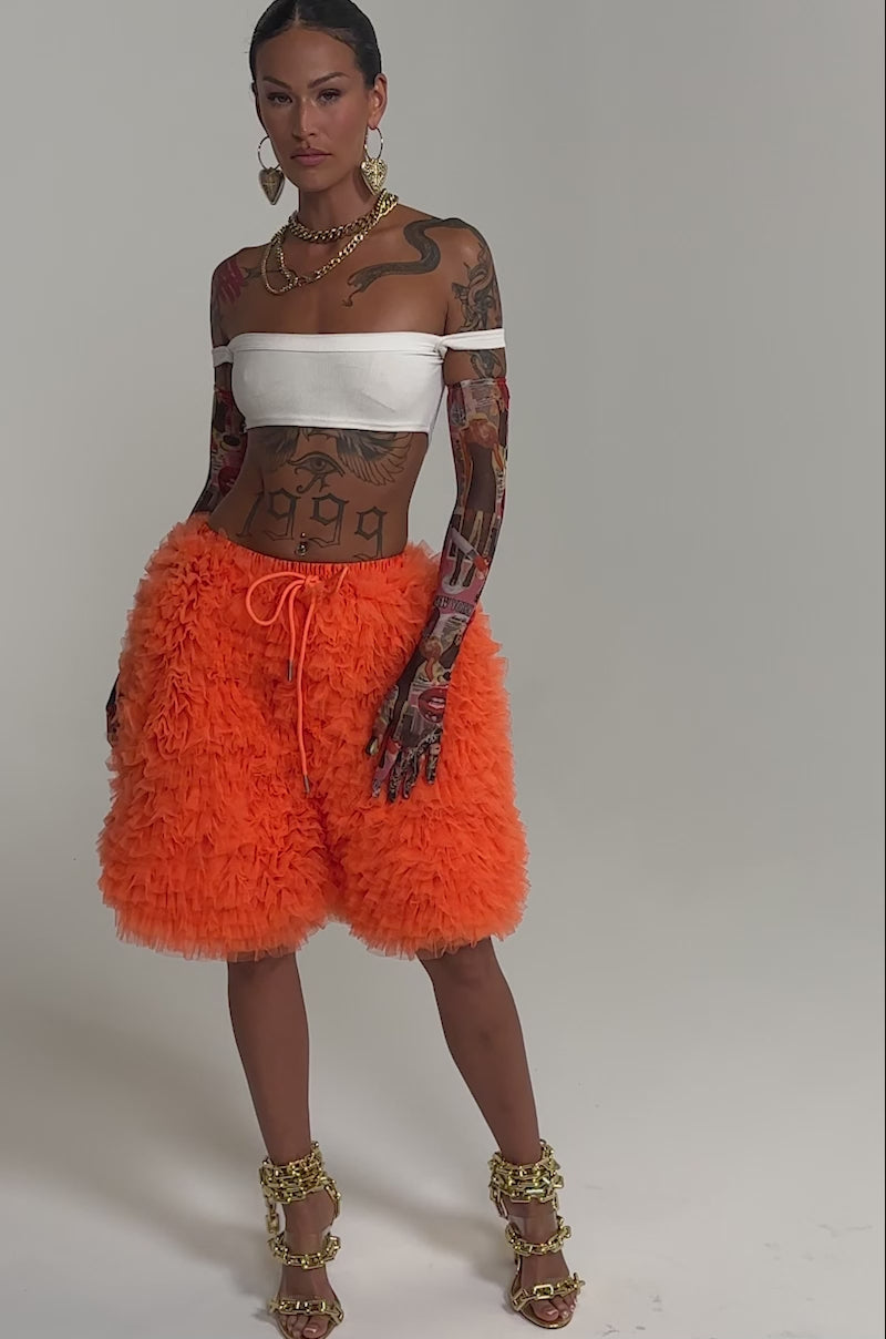 Video of woman modeling the SOMETHING CRAZY RUFFLE SHORT in bright orange, paired with gold high-heeled sandals featuring chain details. 