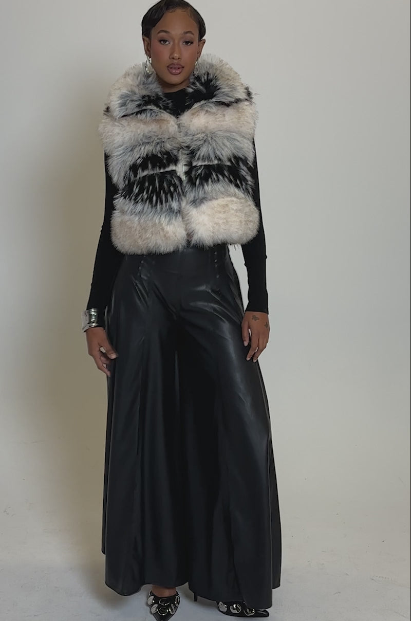 Video of woman modeling the KIKI FAUX FUR VEST—a sleeveless striped faux fur vest with beige and black patterns—styled over a black long-sleeve top and black leather pants, accessorized with a silver cuff bracelet and small earrings.