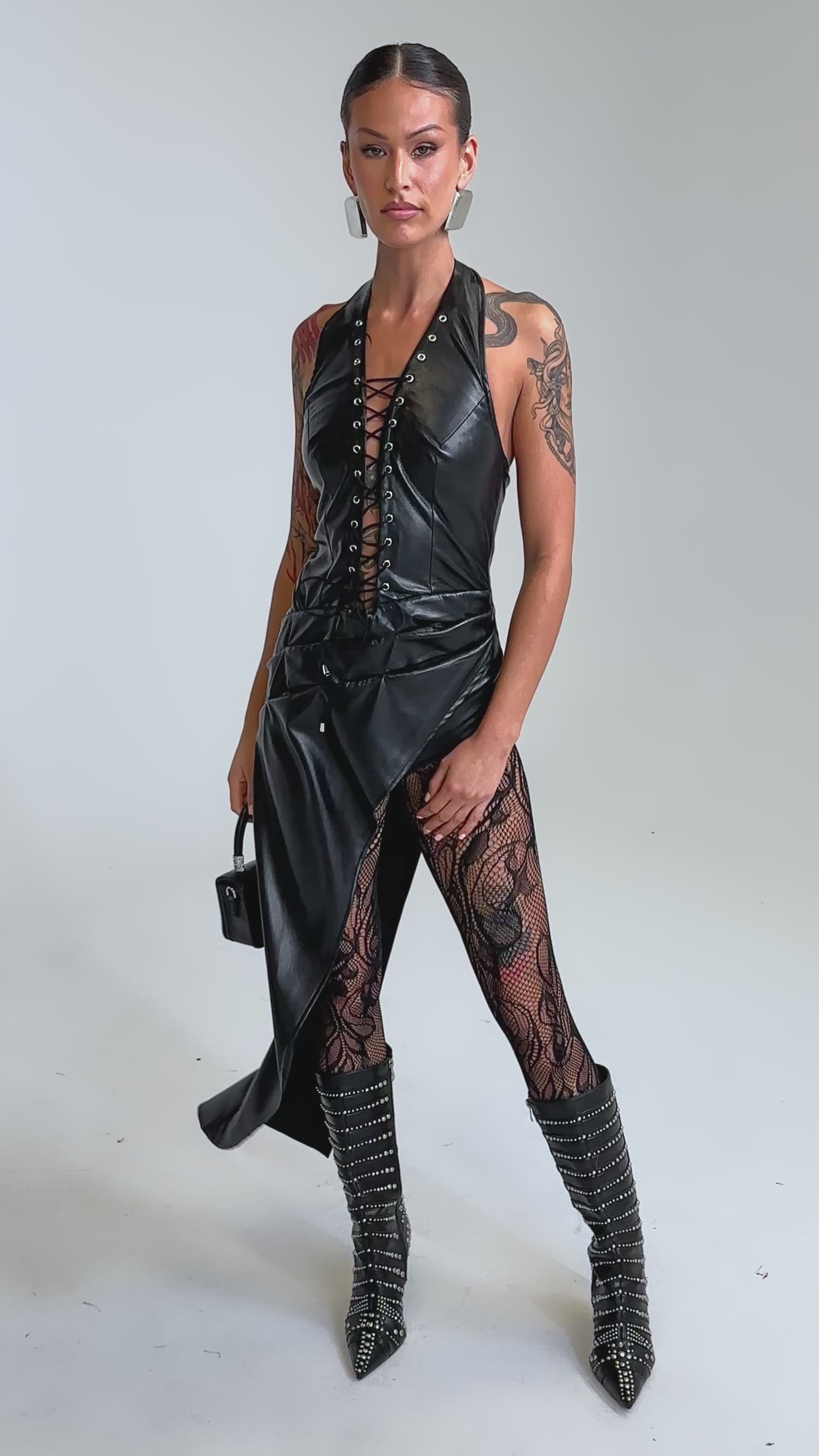 Video of woman modeling the TALIA LACE UP DRAPE TOP and matching skirt, paired with patterned sheer leggings, studded boots, hoop earrings, and a small black handbag.