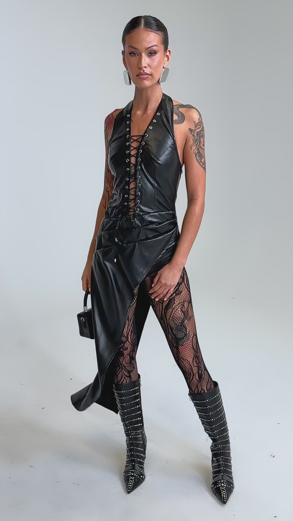 Video of woman modeling the TALIA LACE UP DRAPE TOP and matching skirt, paired with patterned sheer leggings, studded boots, hoop earrings, and a small black handbag.