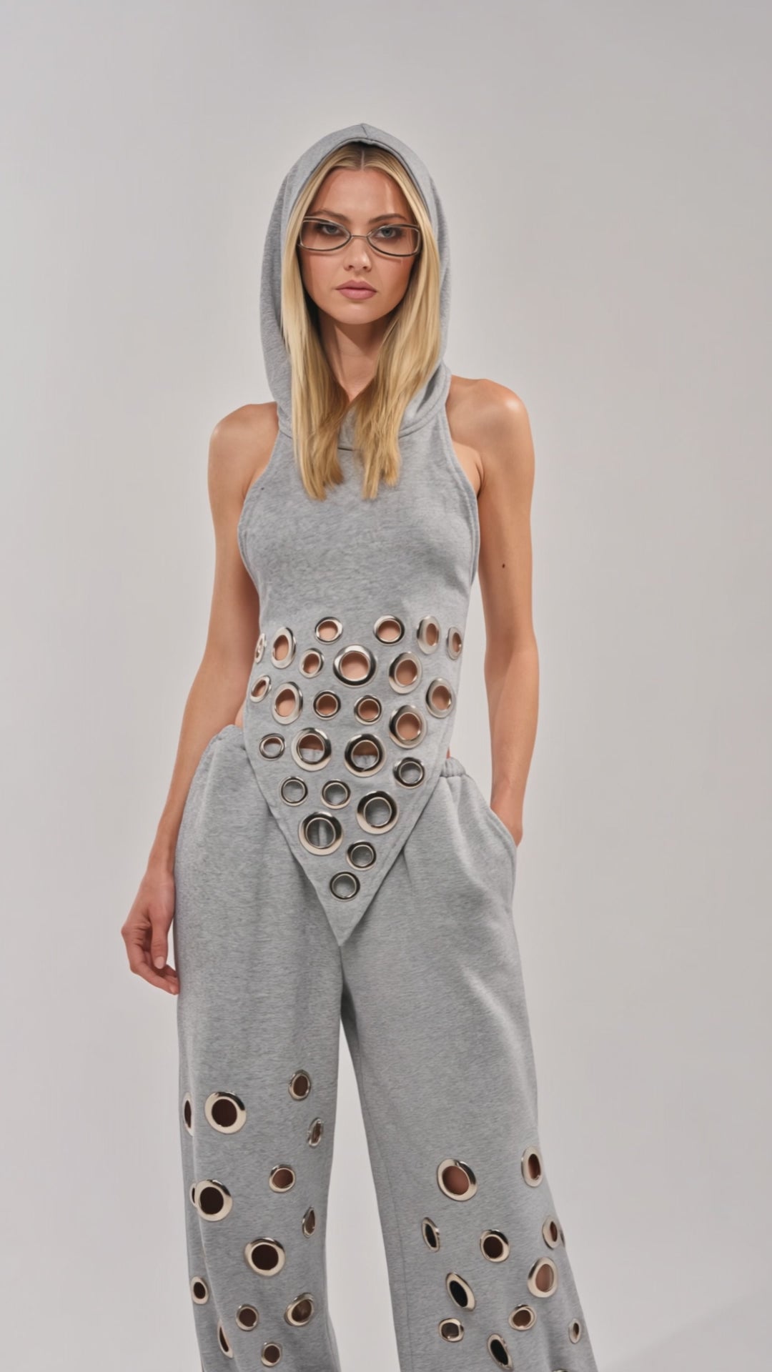 Video of woman modeling the CHECK IT OUT GROMMET TOP—a gray hooded halter with bold metallic grommets—paired with matching gray pants standing against a plain background, exuding streetwear style with straight hair and sunglasses.