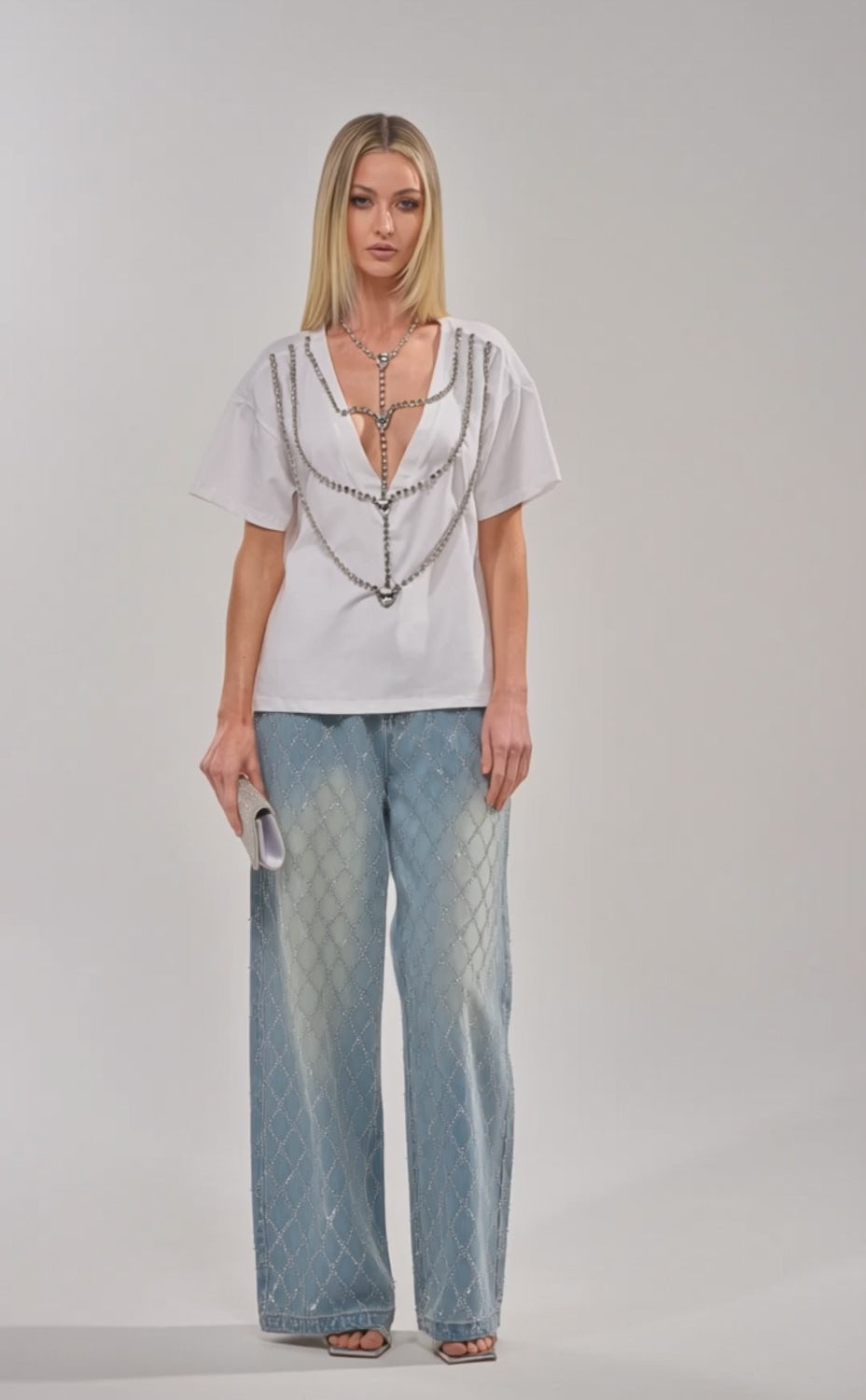 Video of woman modeling the DIAMONDS DANCING RHINESTONE DETAIL TEE, featuring silver chains, paired with diamond-patterned blue jeans. She holds a sparkling clutch and stands against a plain background.