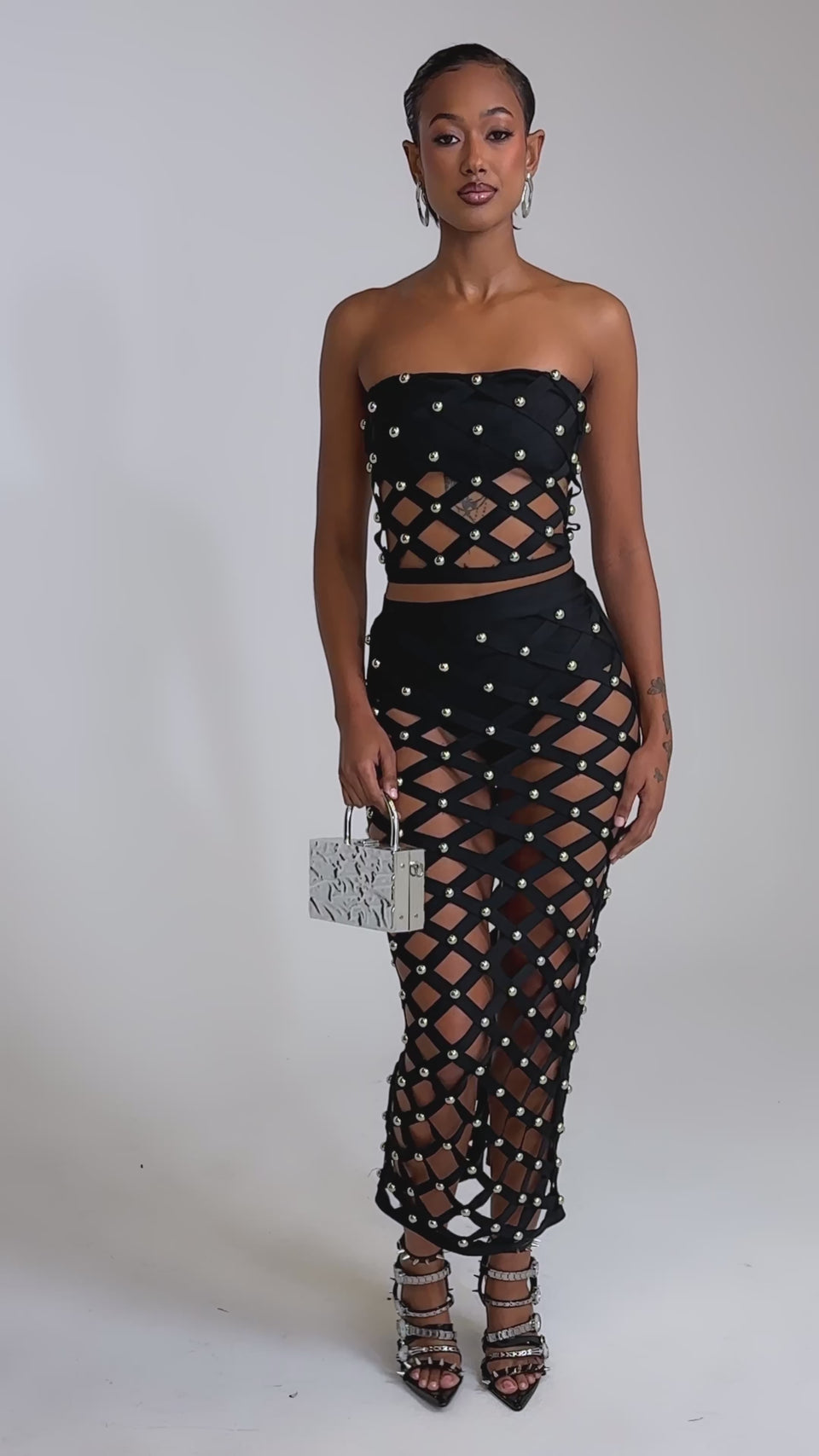 A video of a woman in the CRAZY ENOUGH MAXI SKIRT, a high-waisted, cage-style black skirt with silver studs, paired with a strapless black crop top, strappy heels, and a metallic purse.