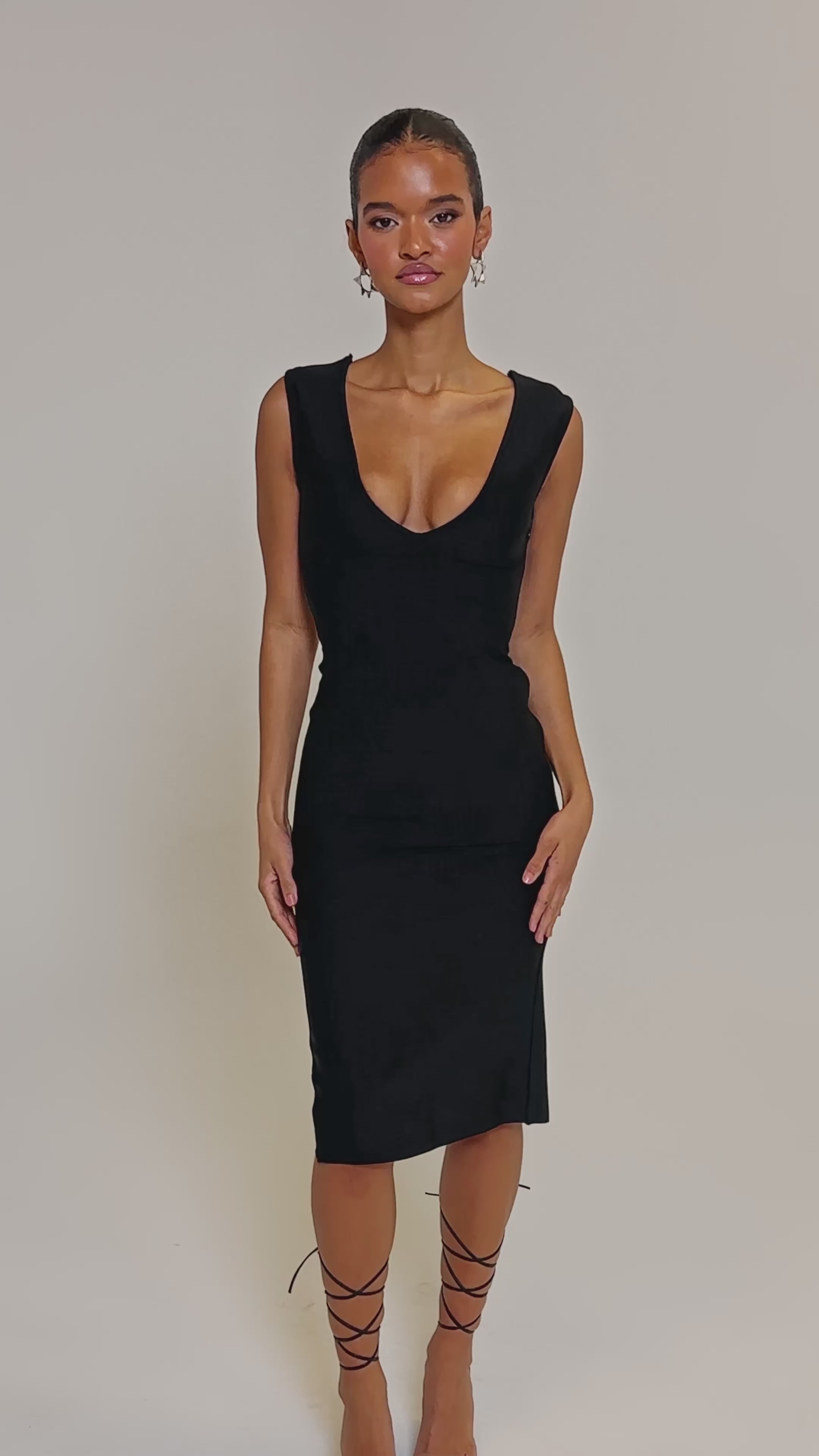 Video of woman modeling the MIDNIGHT LACES CORSET MIDI DRESS, showcasing sleek stretch bandage fabric with lace-up panels on the back and sides. She pairs it with strappy heels and a chic updo, posing against a plain light background.