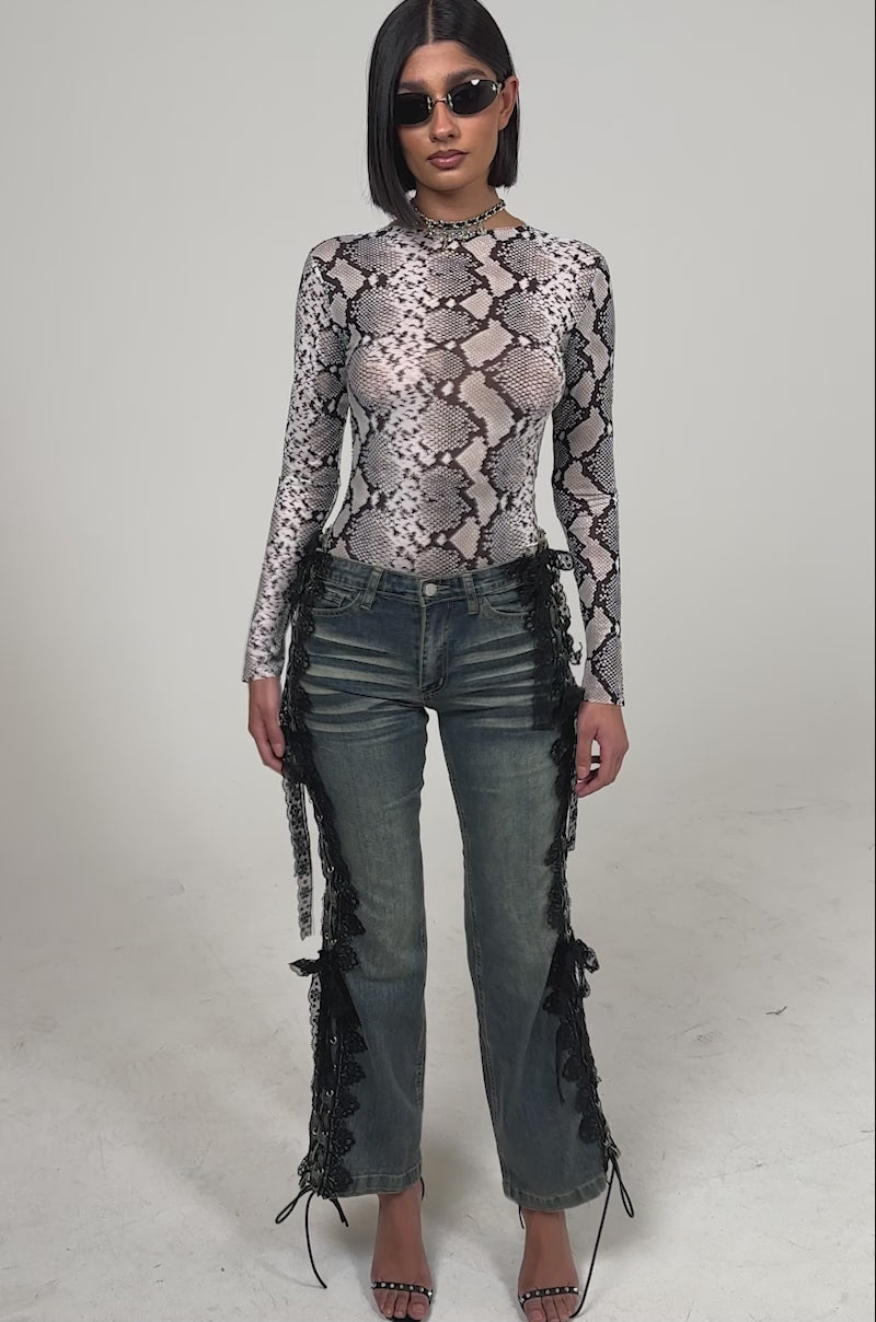 A video of a woman wearing sunglasses, a long-sleeve snakeskin-print top, black strappy heels, and the ELIJAH FLARE LACE DENIM against a plain white background.