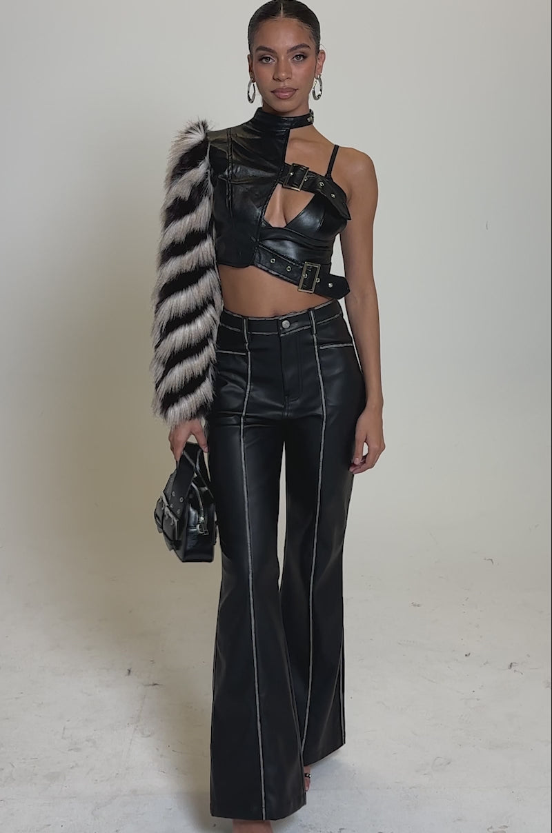 A video of a woman modelling the HIGH AND LOW FUR BLOUSE, a black faux leather one-shoulder crop top with a chic one-sleeve design, paired with high-waisted black flared pants, a striped fur stole, hoop earrings, strappy heels, and a small black handbag.