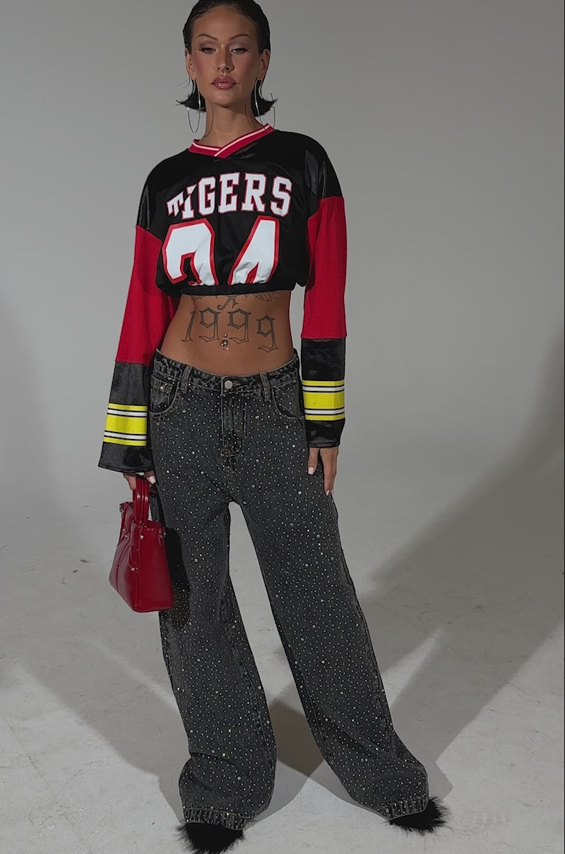 A video of a woman wearing a cropped jersey, RHINESTONE QUEEN EMBELLISHED DENIM, black shoes, hoop earrings, and holds a red handbag. A "1999" tattoo is visible on her stomach.