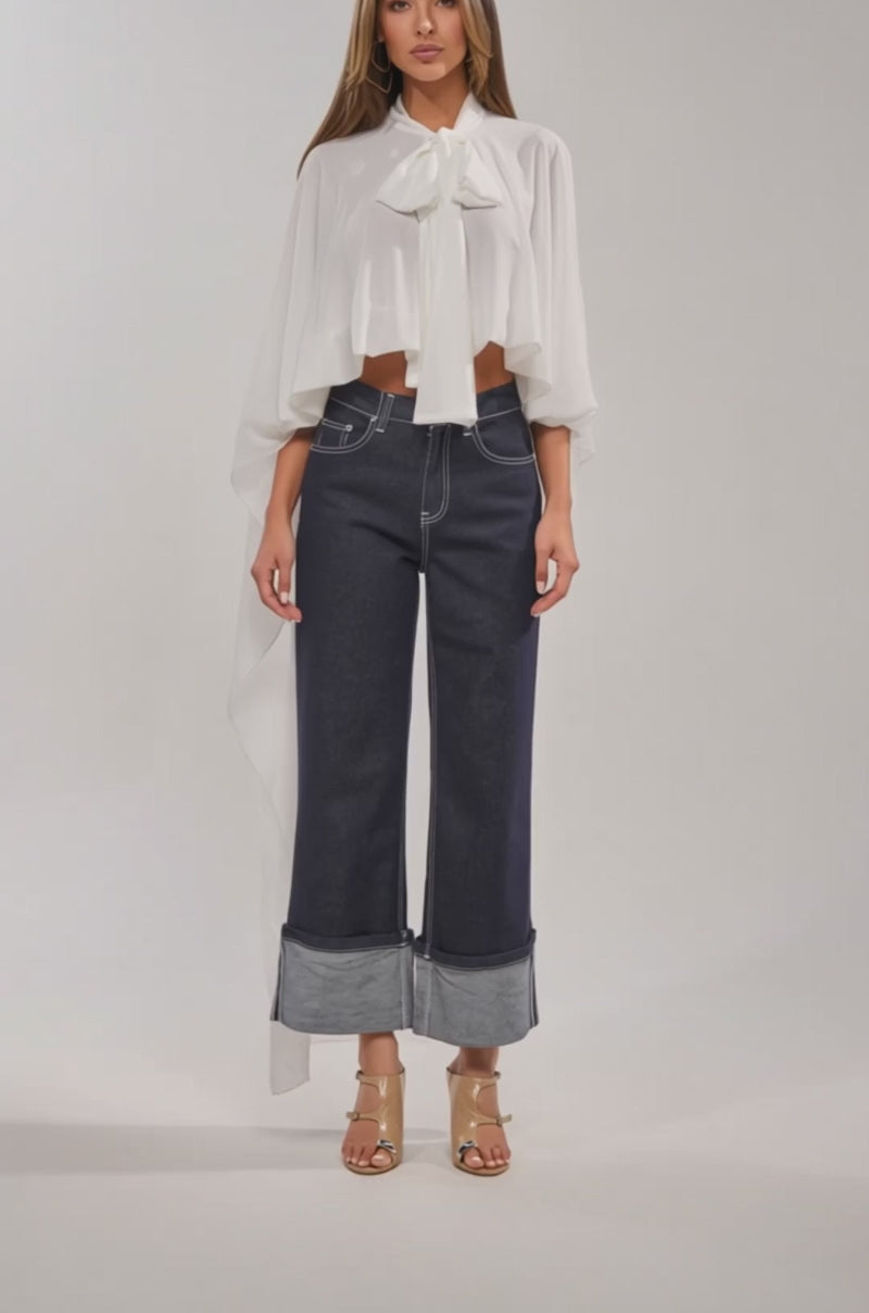 A video of a woman wearing the JENNA CHIFFON BLOUSE with cape-inspired sleeves, paired with wide-leg dark blue cuffed jeans and clear heeled sandals against a plain background.