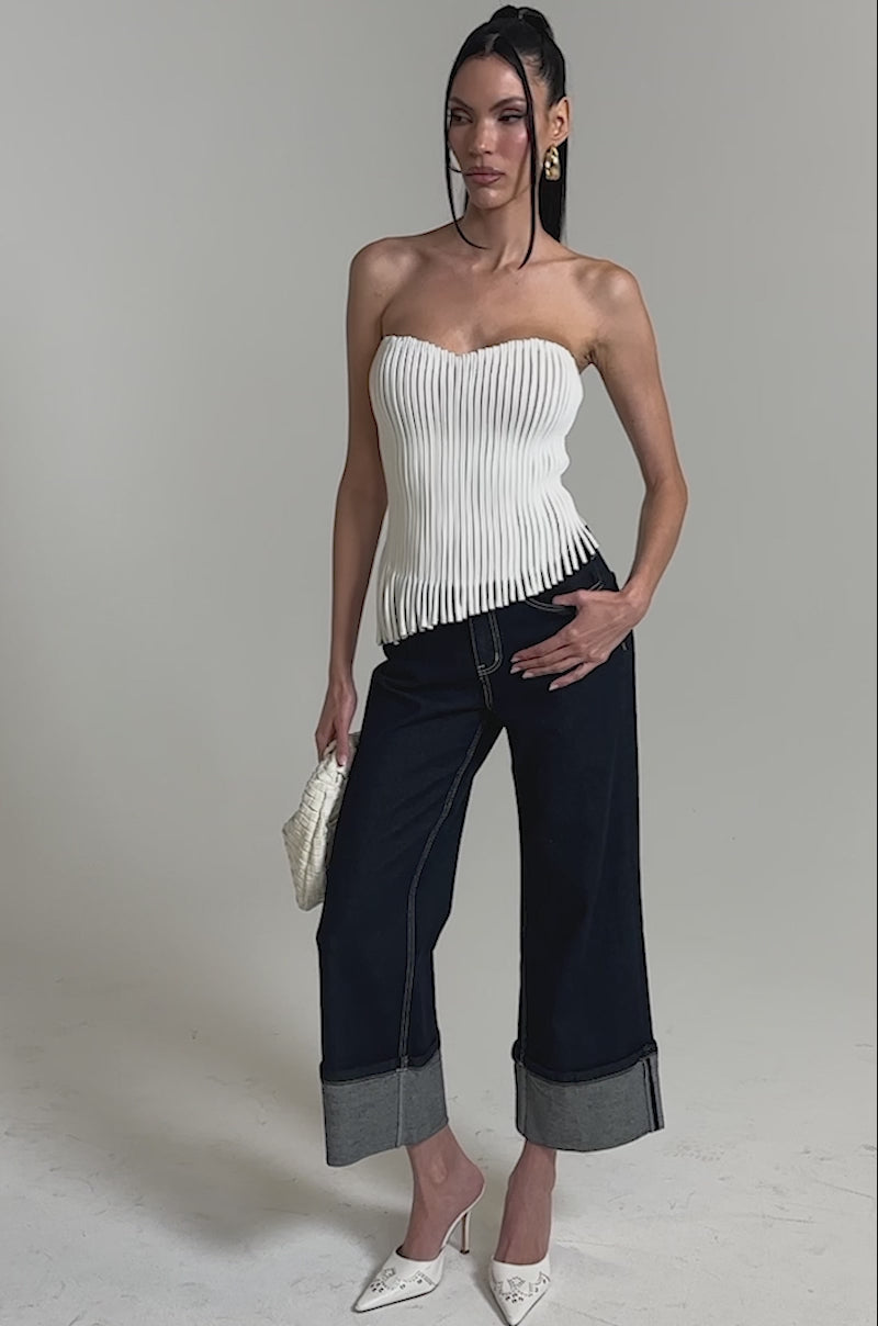 A video of a woman in a studio wearing the white GINA FRINGE CORSET with dark wide-leg cuffed jeans, white pointed-toe heels, and carrying a small white woven handbag.