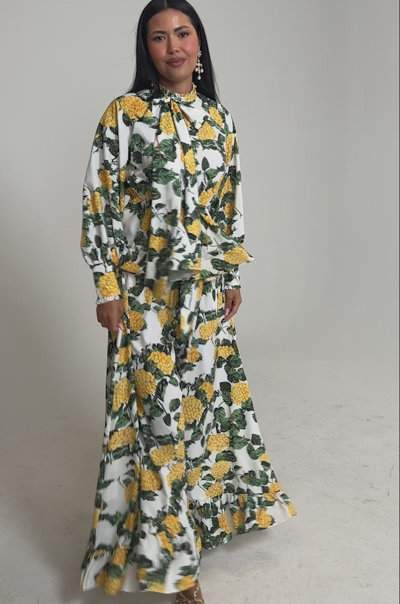 Video of woman modeling the JULIE FLORAL WIDE LEG RUFFLE PANT with a matching long-sleeve tie-neck top. The yellow floral print with green leaves pops against the white base, set on a plain background.