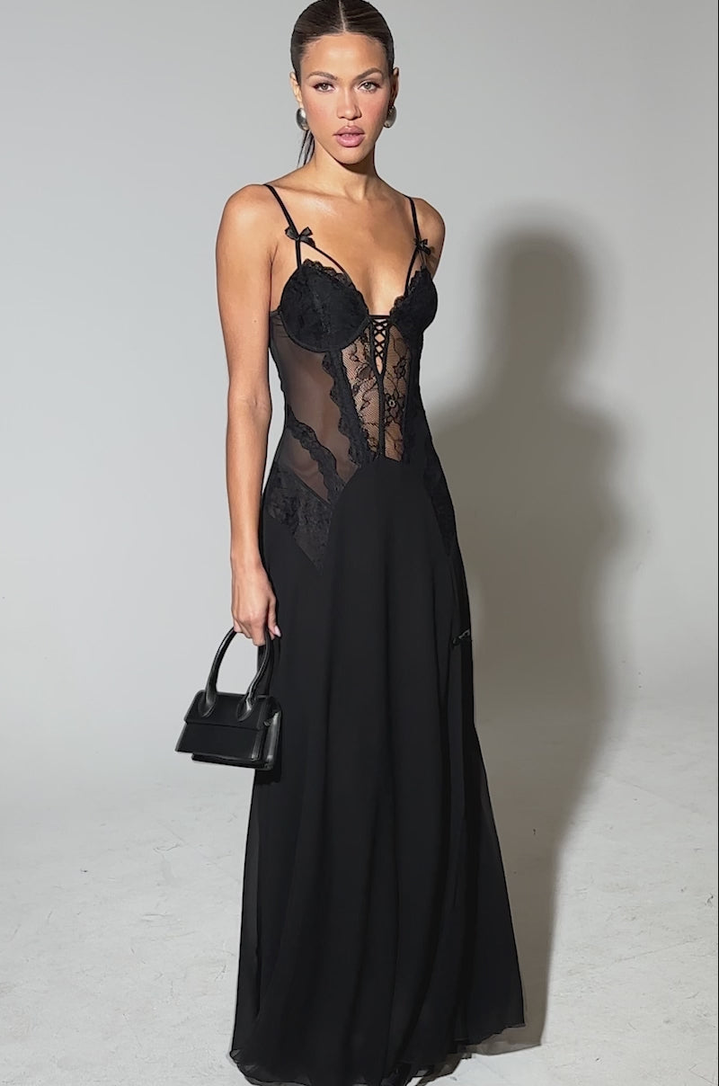 Video of woman modeling the LEILA MAXI DRESS—a long black lace bustier dress with spaghetti straps and a sheer bodice—holds a small black handbag and stands against a plain light background.