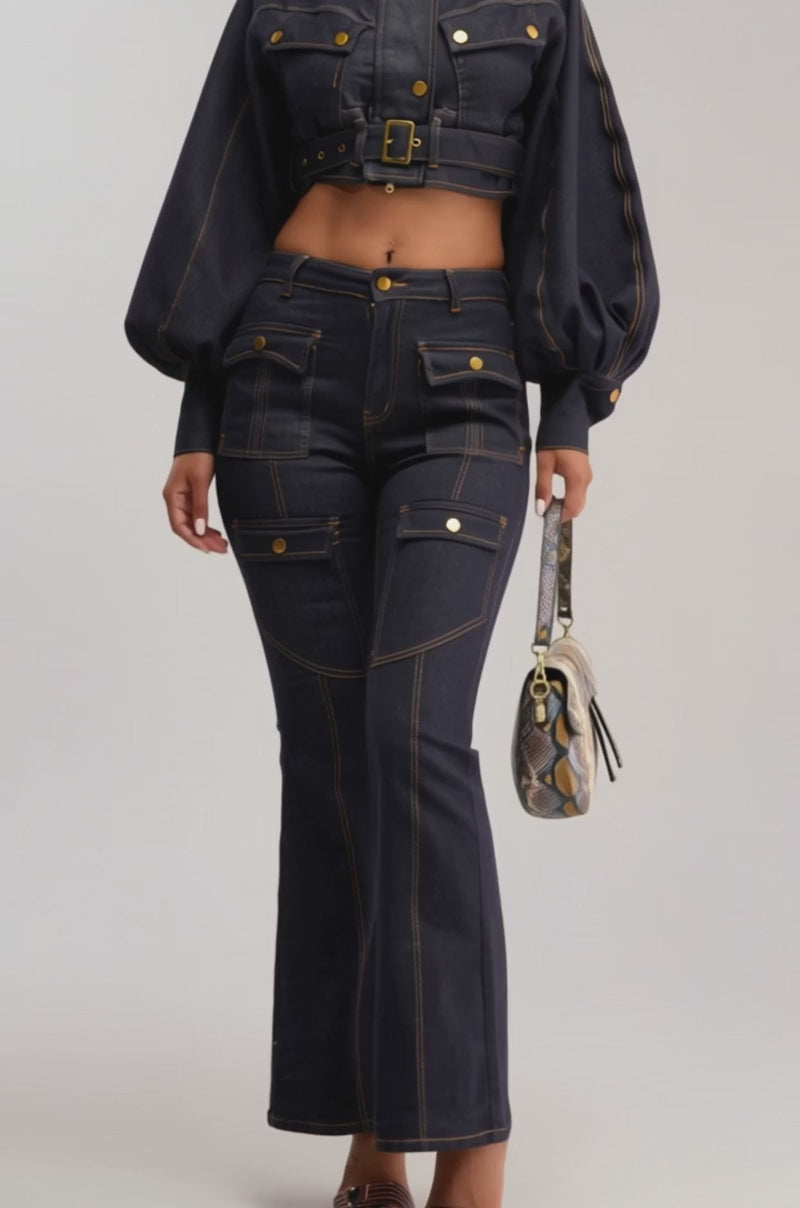 Video of woman modeling the CHIARA DENIM JACKET—a utility-inspired, cropped denim jacket with balloon sleeves, gold buttons, and a belt—paired with matching high-waisted jeans. She carries a small patterned handbag and smiles slightly.