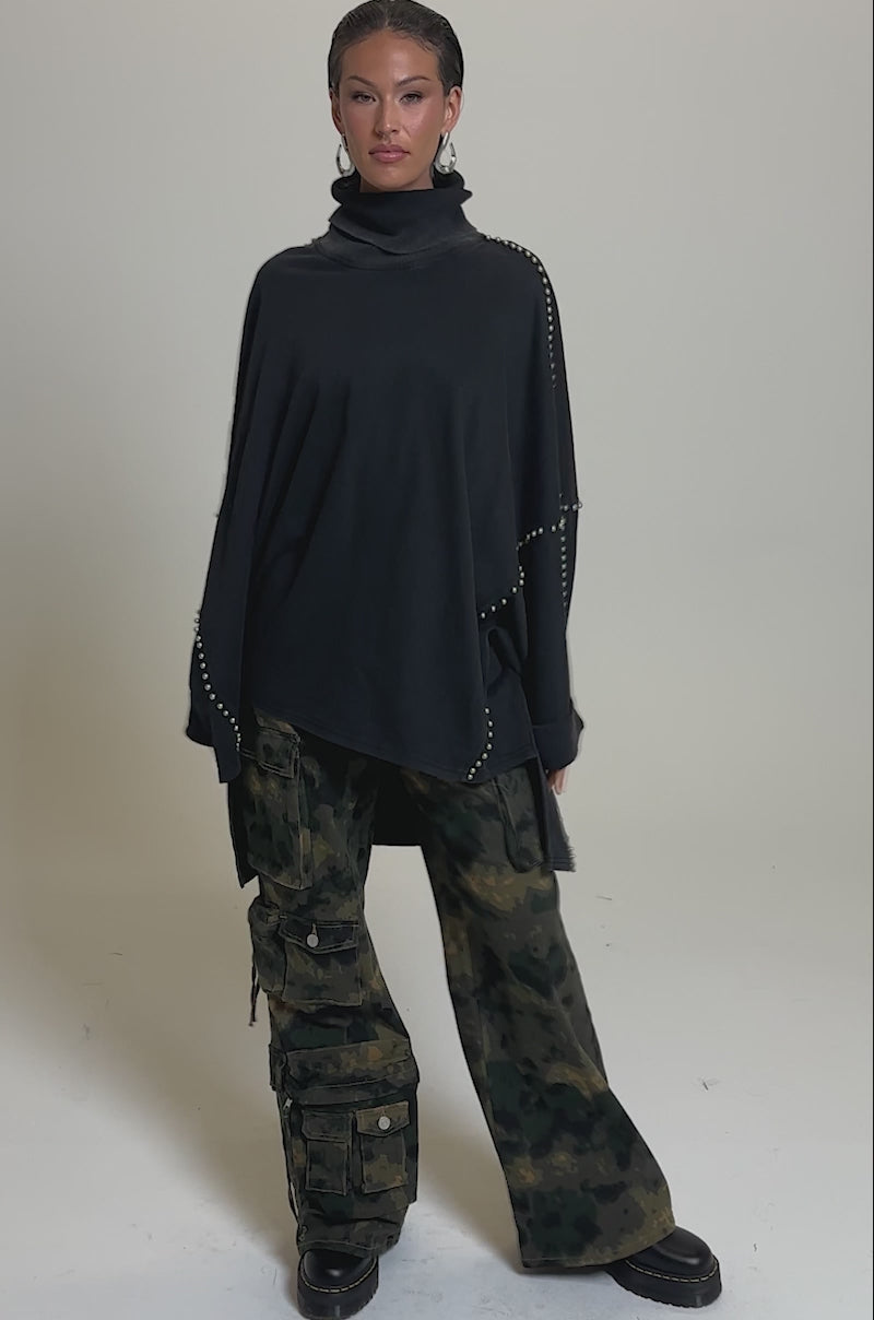 A video of a person wearing the OVER IT TURTLE NECK OVERSIZED SWEATSHIRT with batwing sleeves and studded details, paired with loose camouflage cargo pants and chunky black shoes, against a plain light background.