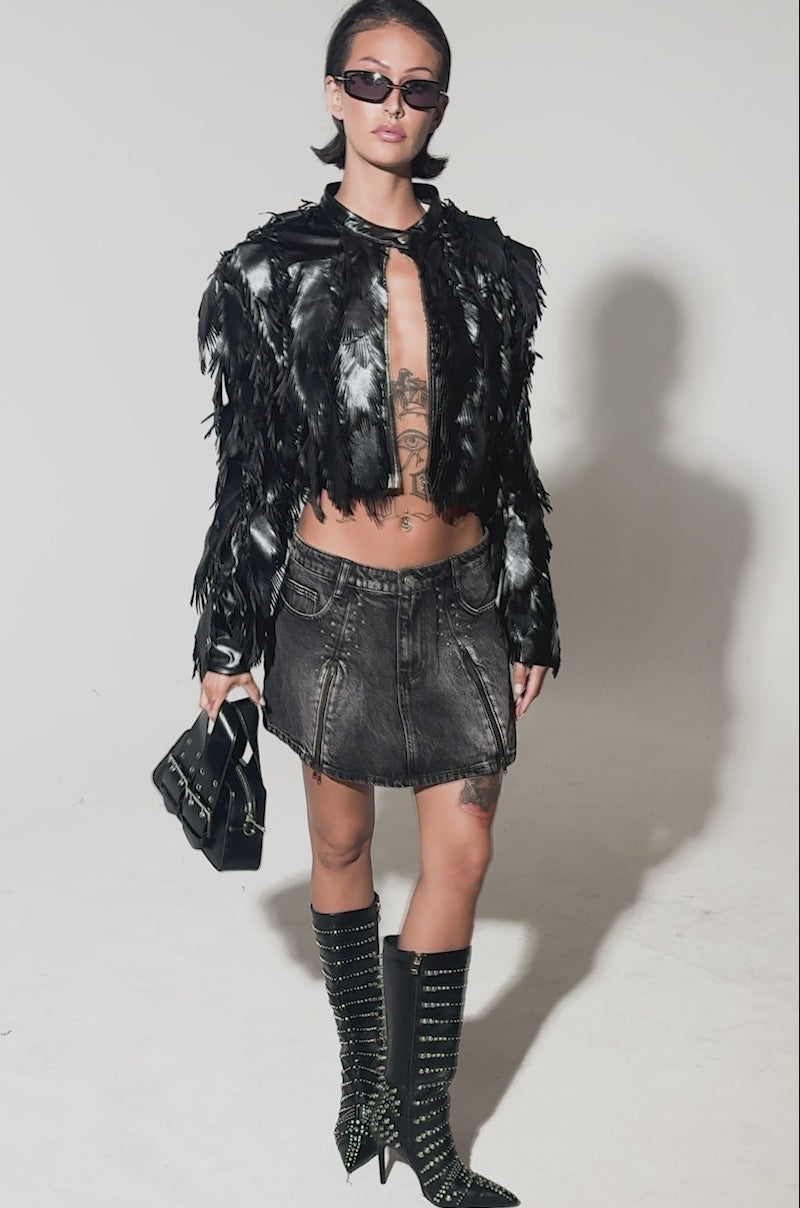 A video of a model wearing sunglasses, the AVANT GARDE LASER CUT BOMBER in black faux leather, a short black denim skirt, studded knee-high boots, and carries a small black handbag. Her torso and thigh tattoos are visible as she stands against a plain background.
