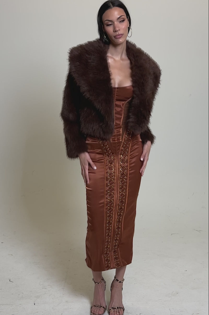 A video of a woman wearing the POPPY Cropped Faux Fur in Brown over a rust satin midi dress with decorative stitching and strappy heels, standing against a plain light gray background.