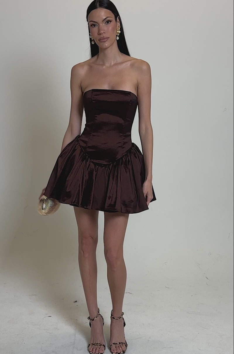 A video of a woman wearing the I LIKE IT LIKE THAT MINI DRESS, a strapless brown mini with a flared bubble skirt. She holds a small textured clutch and pairs statement earrings with strappy high-heeled sandals against a plain backdrop.