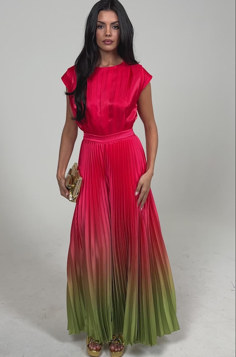 Video of woman modeling the SUNSET VIBES SATIN JUMPSUIT, a sleeveless pleated jumpsuit with a shiny finish that fades from bright red to olive green. She pairs it with a gold clutch and gold platform heels, posing against a plain background.