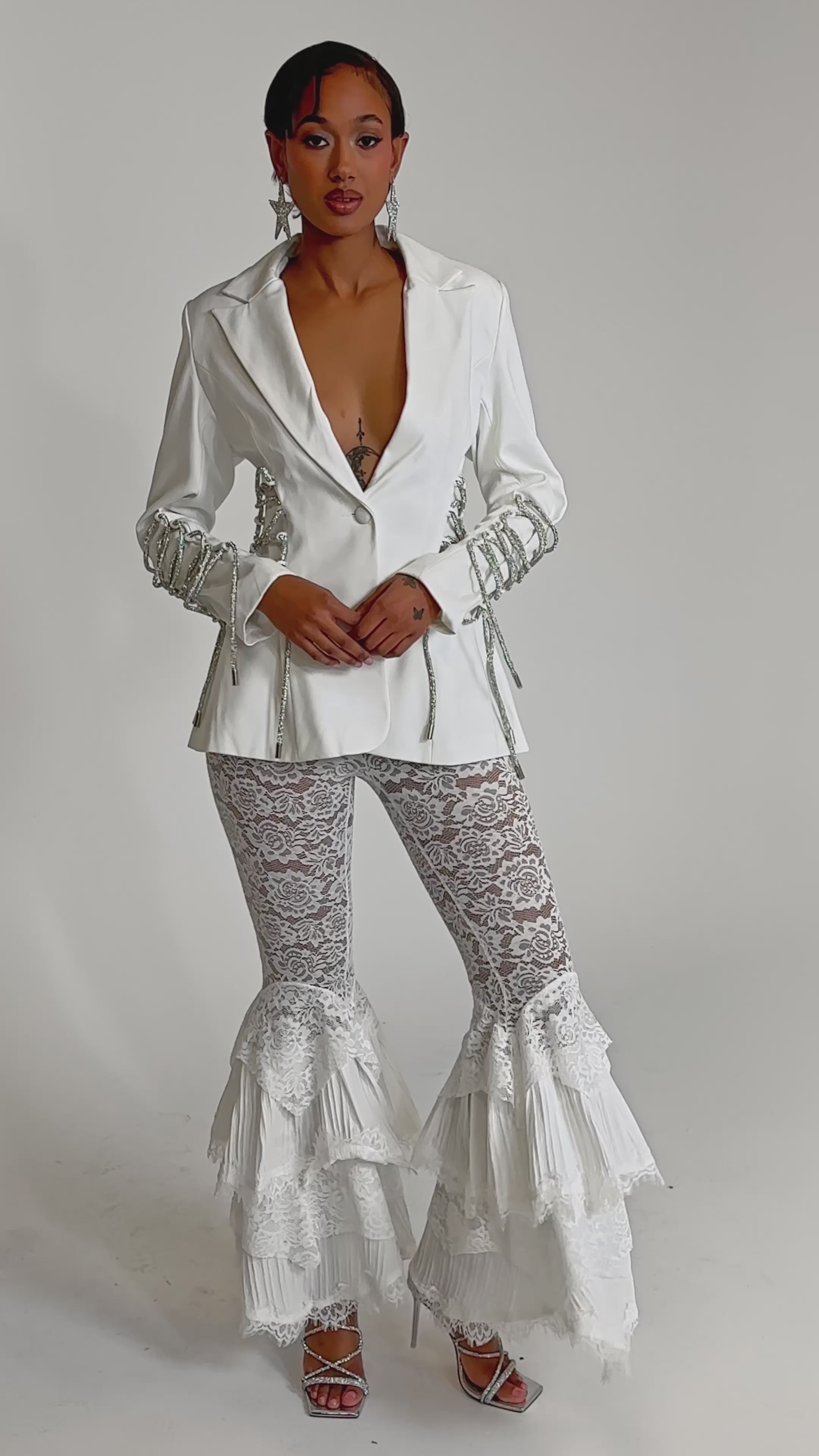A video of a woman wearing the PRETTY PLEASE LACE UP BLAZER, featuring a structured white silhouette with silver chain details, paired with white lace flared pants, statement star earrings, and silver heels against a plain light backdrop.