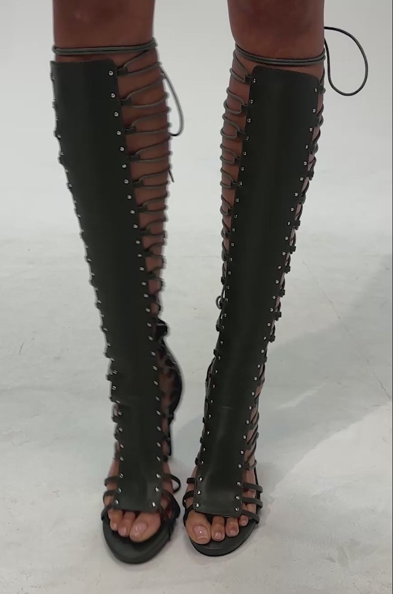 Video of woman modeling the AZALEA WANG ROSALINA OLIVE GLADIATOR TO THE KNEE SANDAL—knee-high gladiator sandals in dark olive green, featuring lace-up details with metal eyelets and stiletto heels, for an alluring boho-chic look.