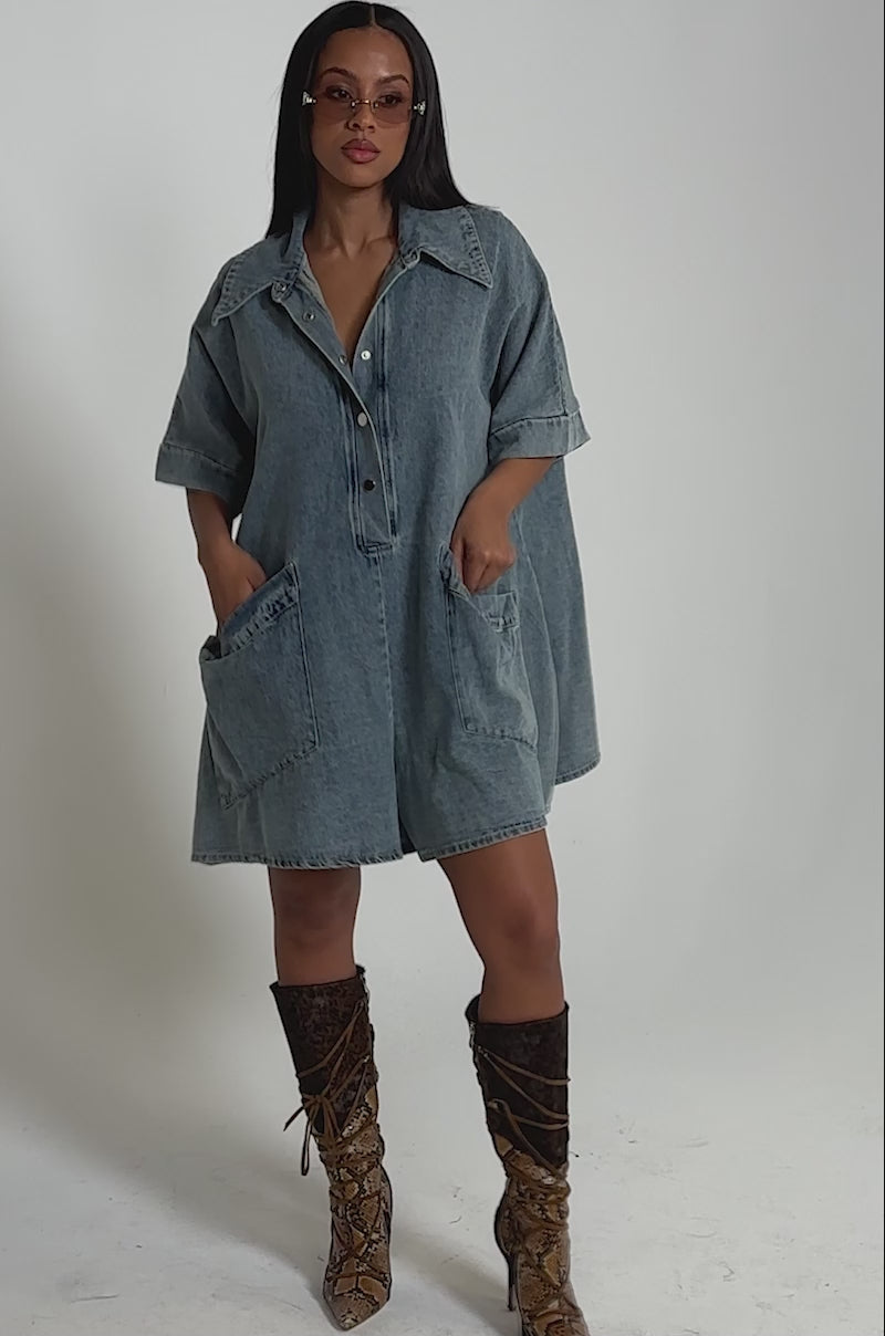 A video of a woman in the MELODY COLLARED DENIM ROMPER with large pockets and snap buttons, styled with knee-high brown snakeskin lace-up boots and sunglasses.