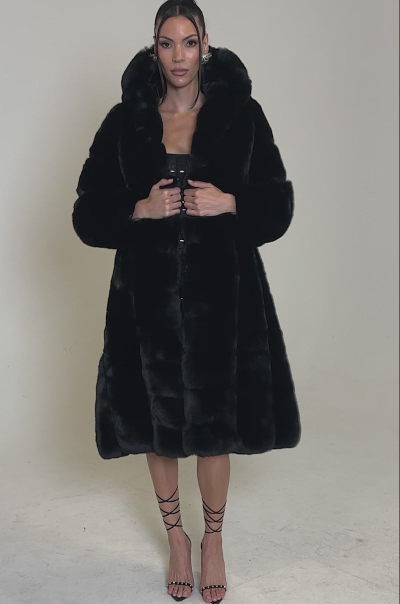A video of a woman standing against a plain background, wearing the BERLIN HOODED FAUX FUR IN BLACK with a large collar, strappy black heels, and her hair pulled back.