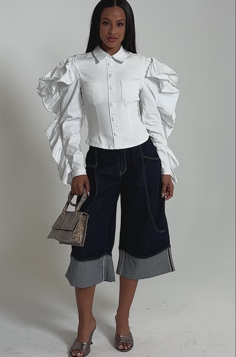 A video of a woman wearing the DREA RUFFLE SLEEVE BUTTON DOWN in white with wide-leg dark denim capris, open-toed heels, and a woven handbag.