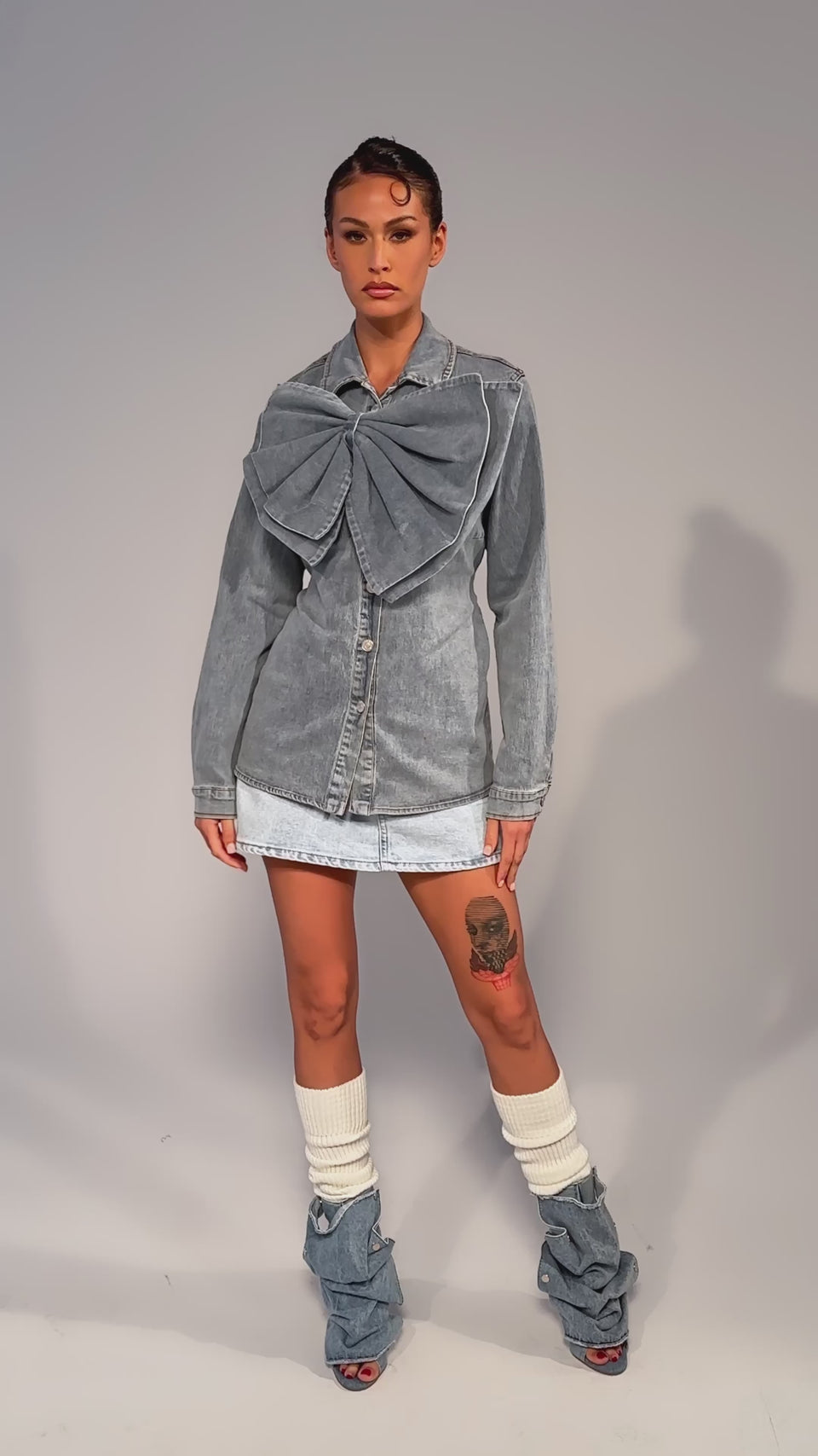 Video of woman modeling the TWO-TONE BOW DENIM BUTTON DOWN SHIRT, featuring an oversized denim bow on one shoulder. Her hair is up, she wears a chunky brown bracelet, and stands before a plain gray background.