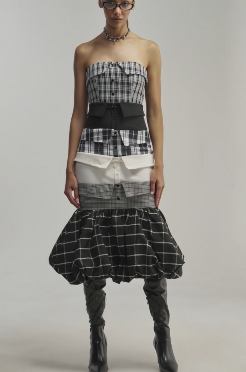 Video of a woman modeling the CHANGING IT UP PLAID MIDI DRESS. This strapless midi dress features a bubble hemline, while its layered skirt—crafted from an assortment of black, white, and plaid fabrics.