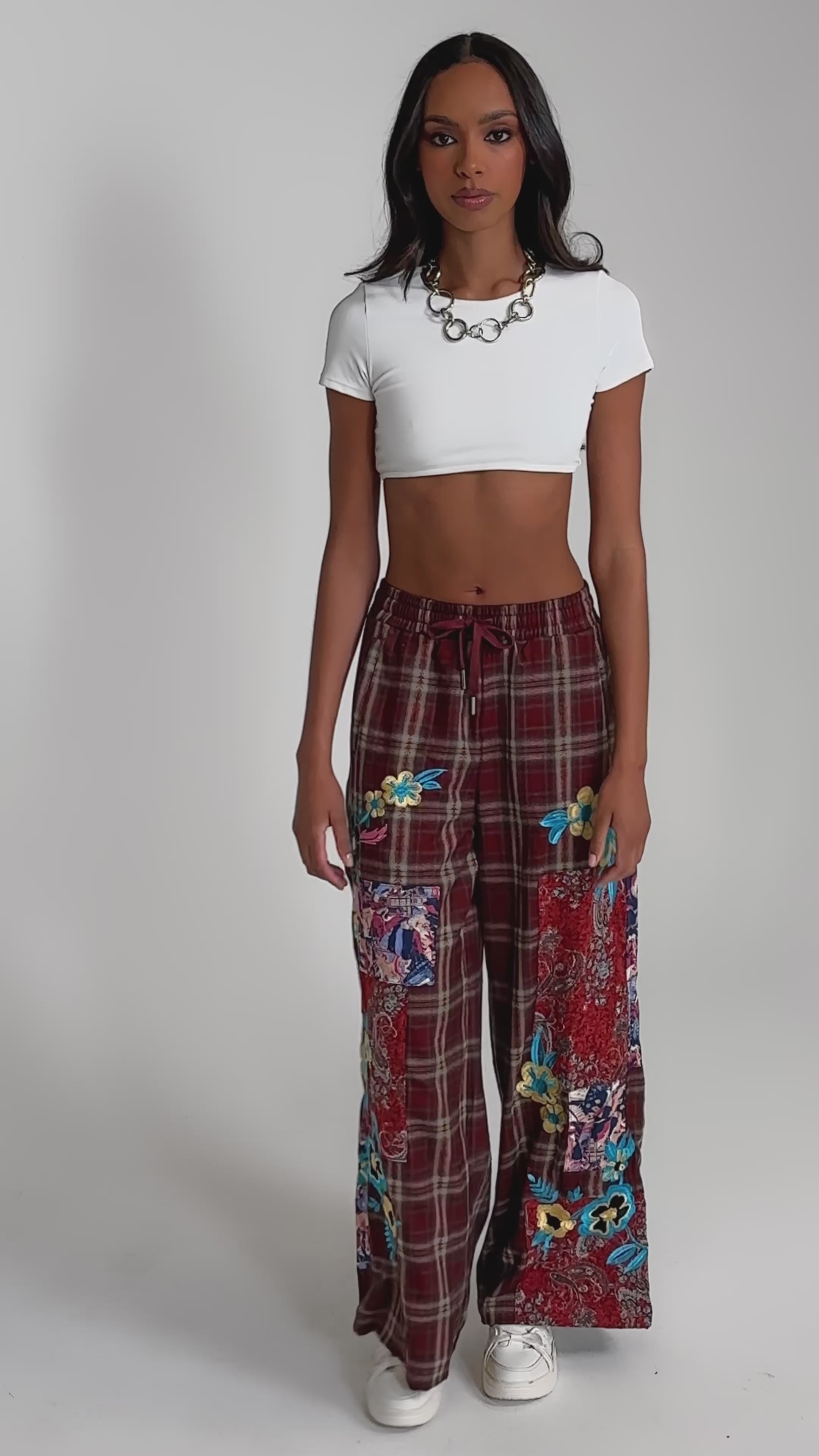 Video of woman modeling the KRIYSTA FLORAL PATCH PLAID PANT—wide-leg maroon plaid pants with colorful floral patchwork—paired with a white cropped t-shirt, white shoes, and a chunky chain necklace against a plain background.