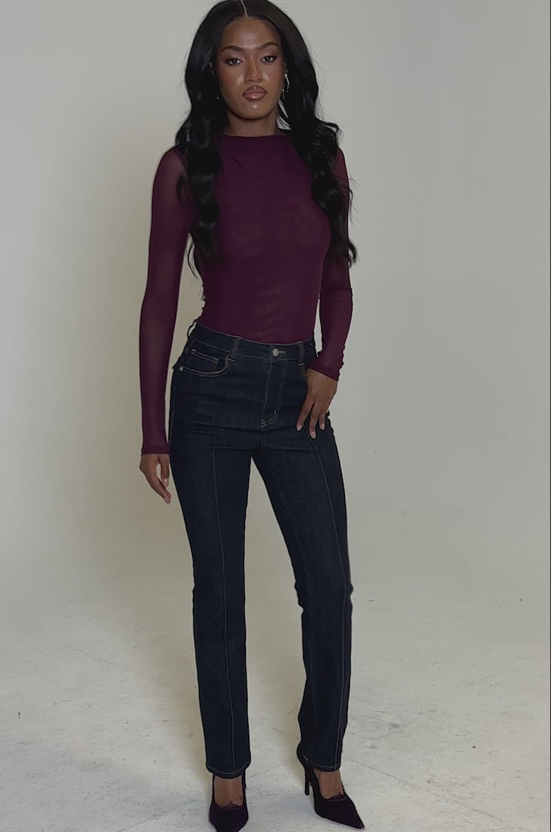 A video of a woman wearing the PAXTON Sexy Mesh Long Sleeve Bodysuit in plum purple, styled with dark blue high-waisted jeans and burgundy heels, her long wavy hair flowing against a neutral background.