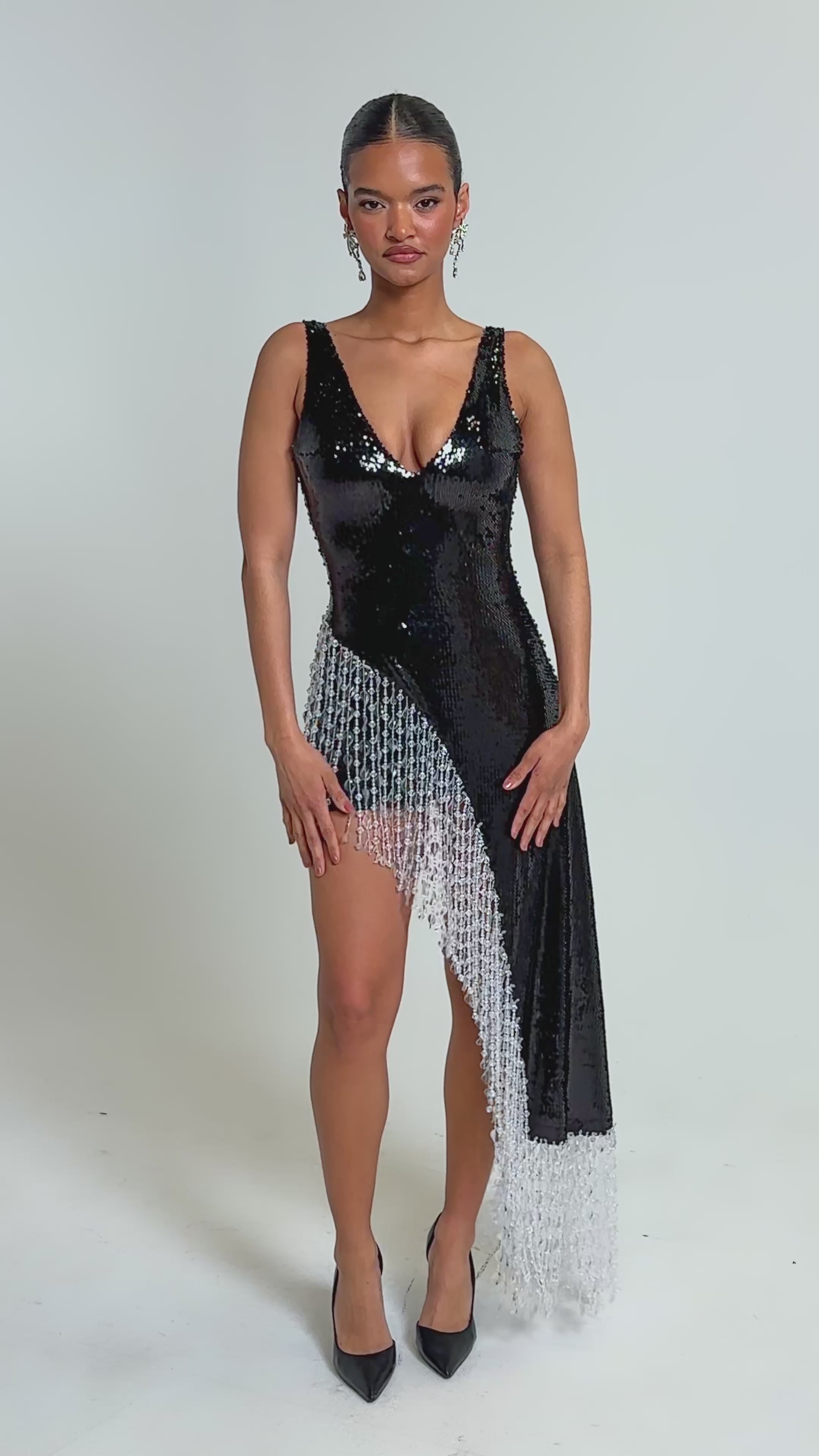 Video of woman modeling the NEW MONEY SEQUIN BLOUSE, a sleeveless black sequin top with an asymmetrical hem and long white beaded fringe trim. She pairs it with black high heels and statement earrings.