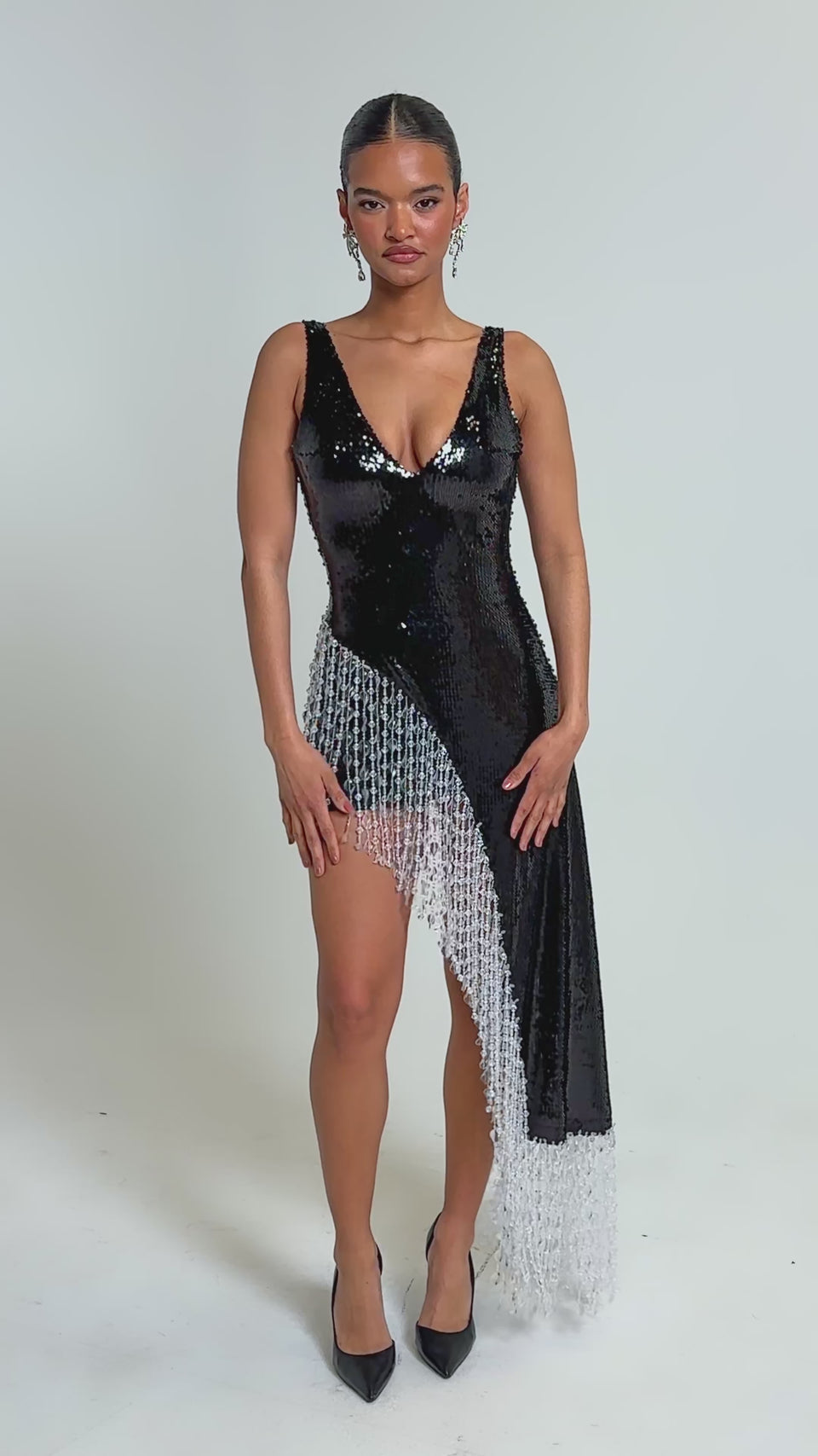Video of woman modeling the NEW MONEY SEQUIN BLOUSE, a sleeveless black sequin top with an asymmetrical hem and long white beaded fringe trim. She pairs it with black high heels and statement earrings.