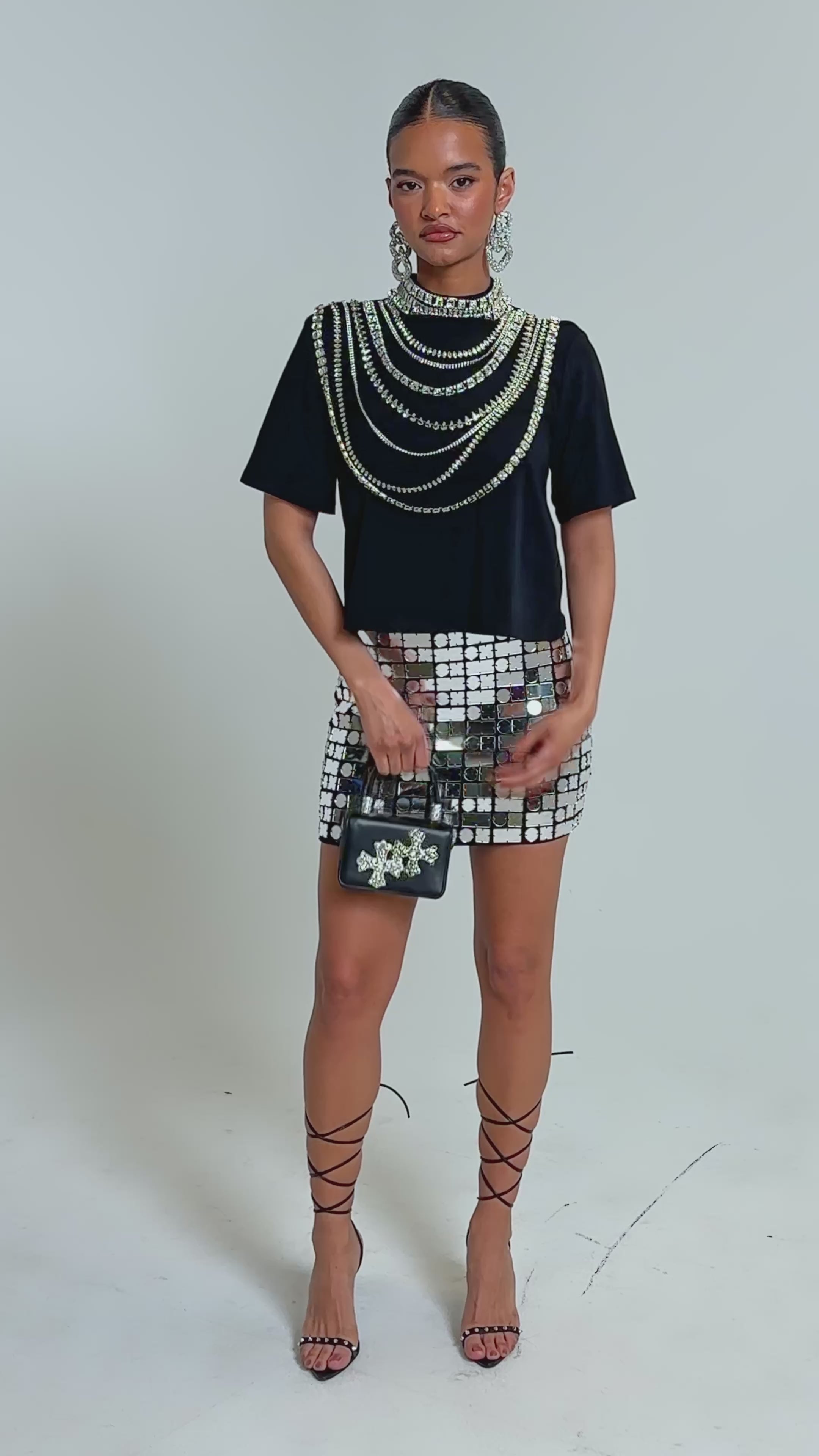 Video of woman modeling the ROYALTY RHINESTONE TEE with layered silver statement necklaces, bold earrings, and a shiny skirt adorned with silver square mirrors. Her hair is neatly slicked back.