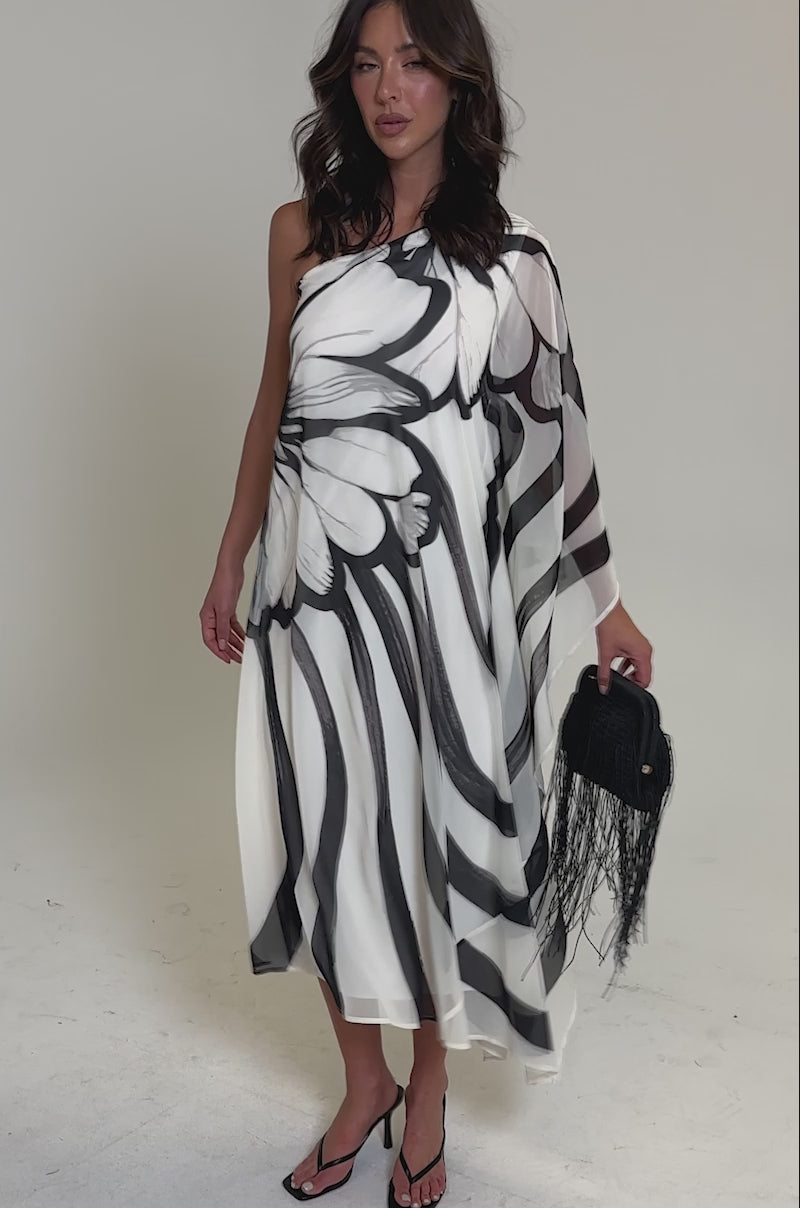 Video of woman modeling the TROPICAL NIGHTS ONE SHOULDER MAXI DRESS with bold black abstract florals, black heeled sandals, and a black fringed handbag