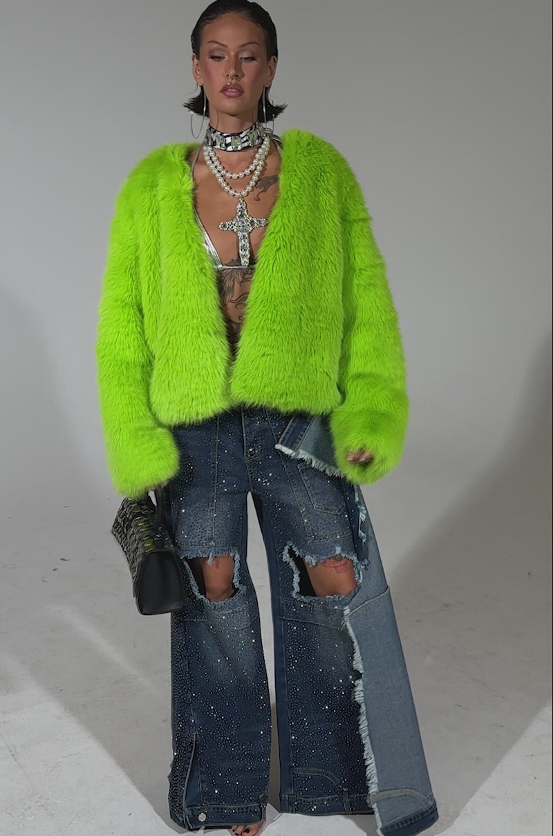A video of a woman confidently posing against a plain background wearing the SLIME TIME FAUX FUR COAT IN LIME, paired with distressed wide-leg embellished jeans, layered necklaces, and holding a studded handbag.