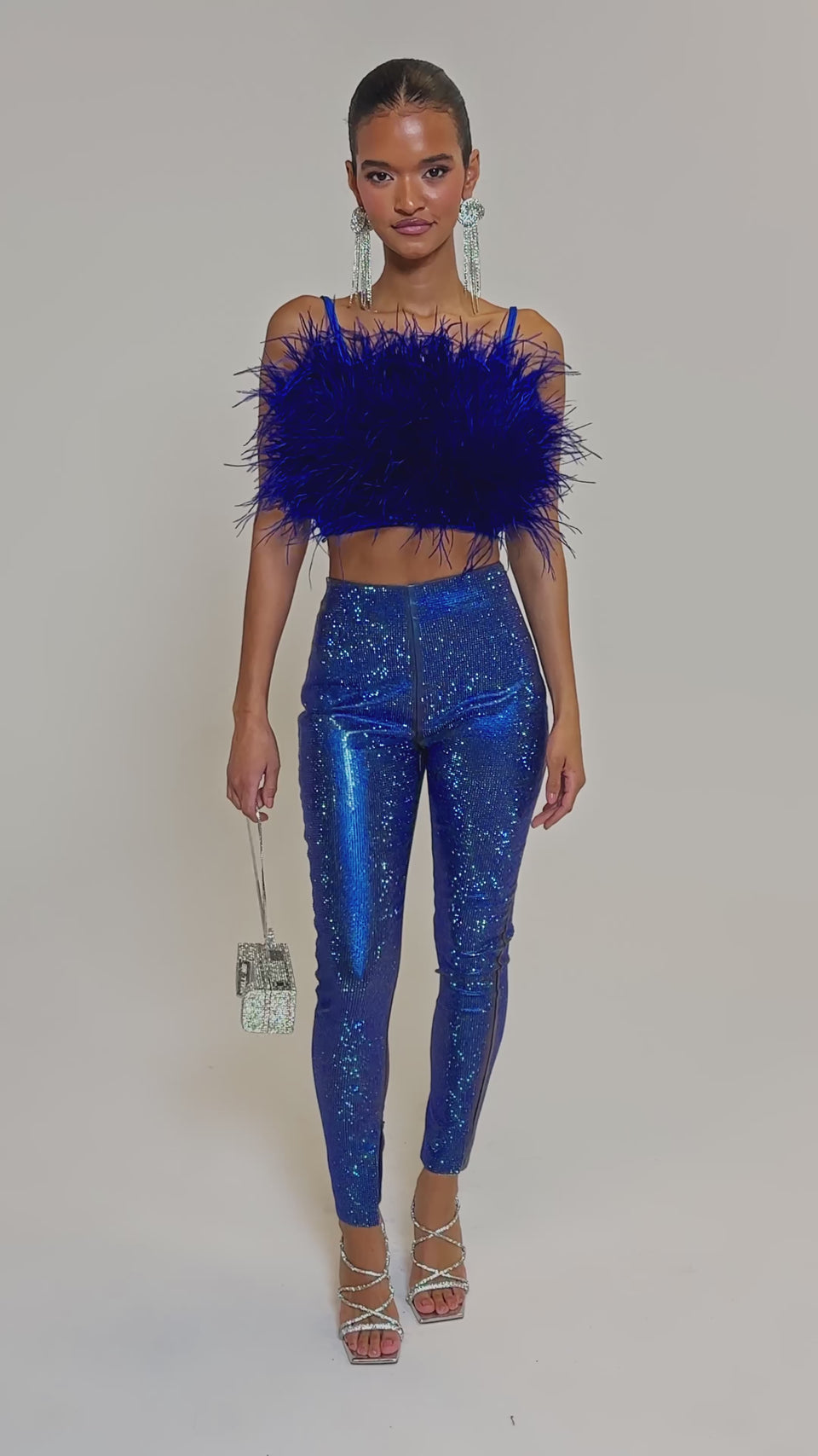 Video of woman modeling the HOTTIE ON THE BLOCK SEQUIN LEGGING in blue with a cobalt feathered crop top, holding a silver handbag and wearing strappy heels against a light gray background.